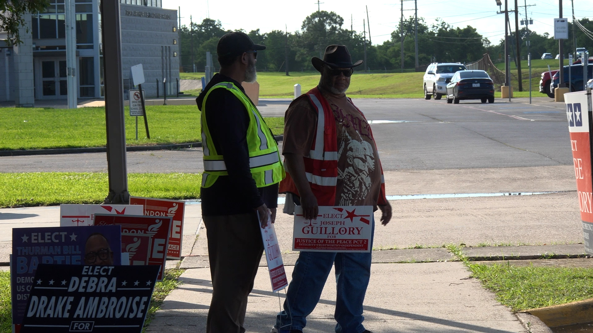 Two men in reflective vests standing by political campaign signs outside a building.