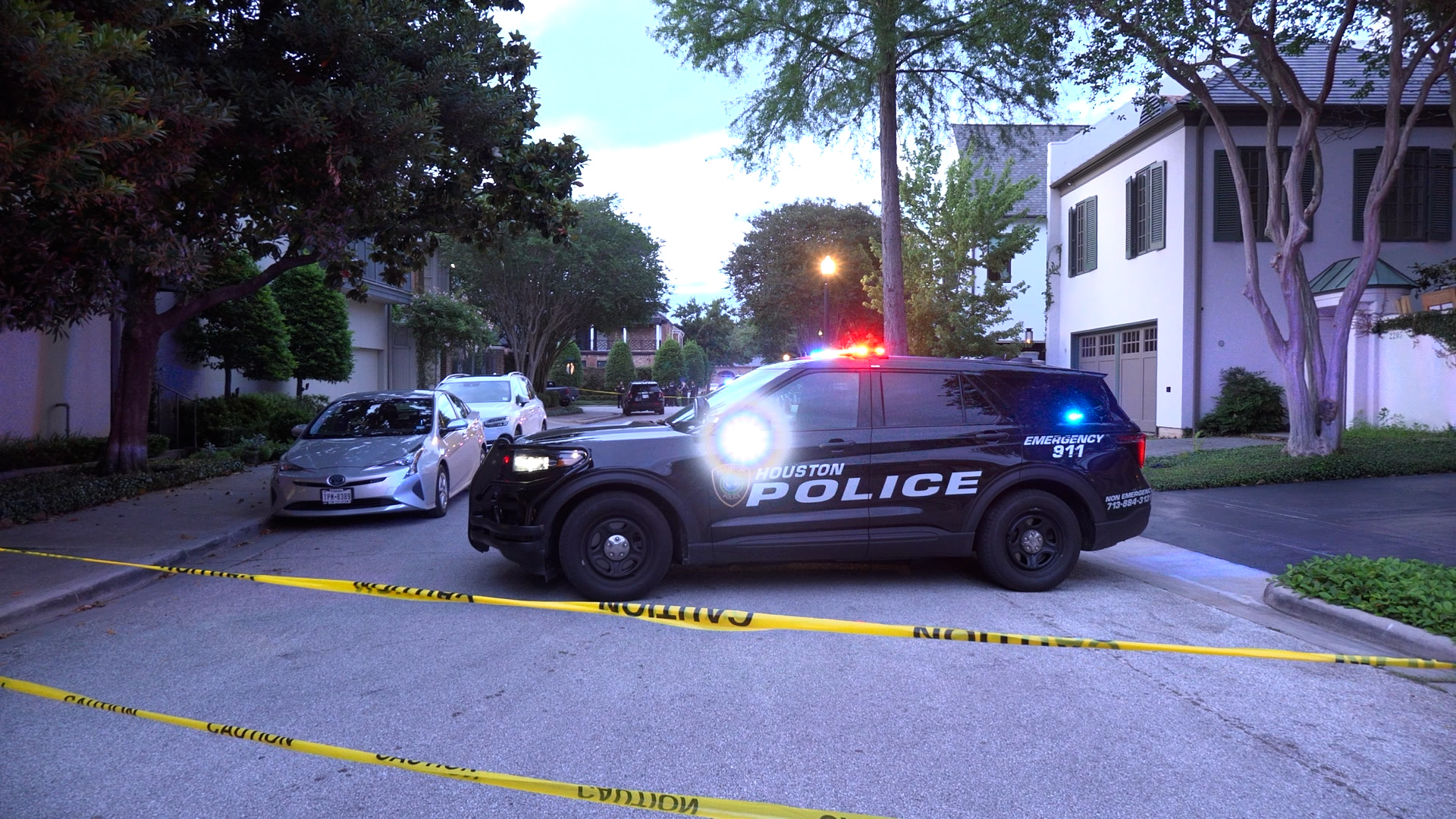 Houston Police SUV with red/blue lights on a residential street, crime scene tape cordoning off the area.