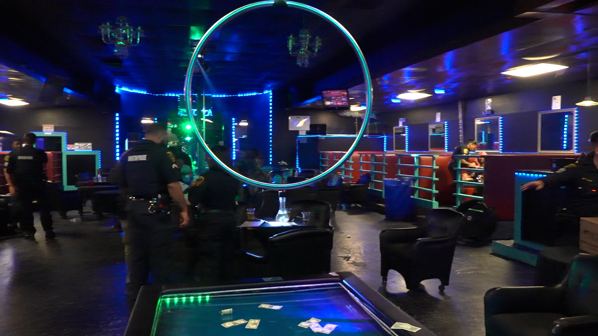 Interior of a neon-lit arcade or entertainment venue with a large circular illuminated hoop in the center and blue LED lighting along walls; people stand near game stations and seating areas in the background.