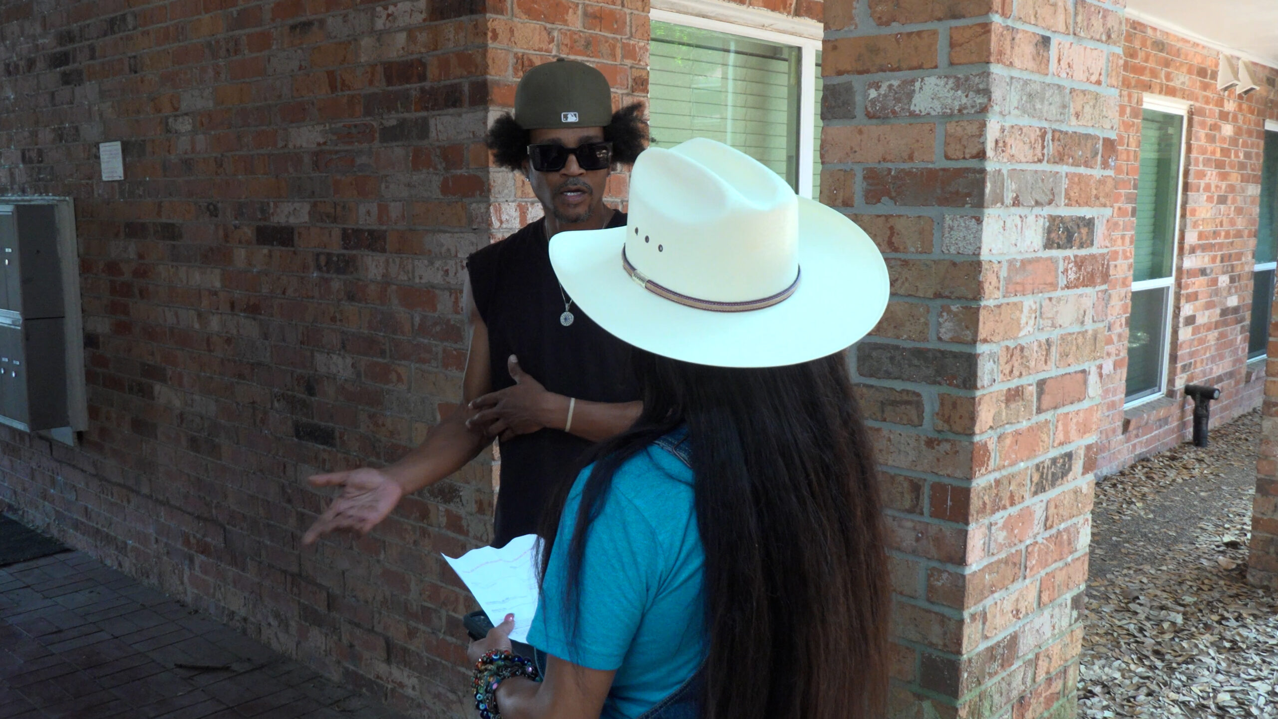 Two people conversing outside a brick building: a woman with long dark hair in a turquoise shirt wearing a large white hat holds papers, facing away from the camera, while a man wearing sunglasses and a green cap gestures toward her.