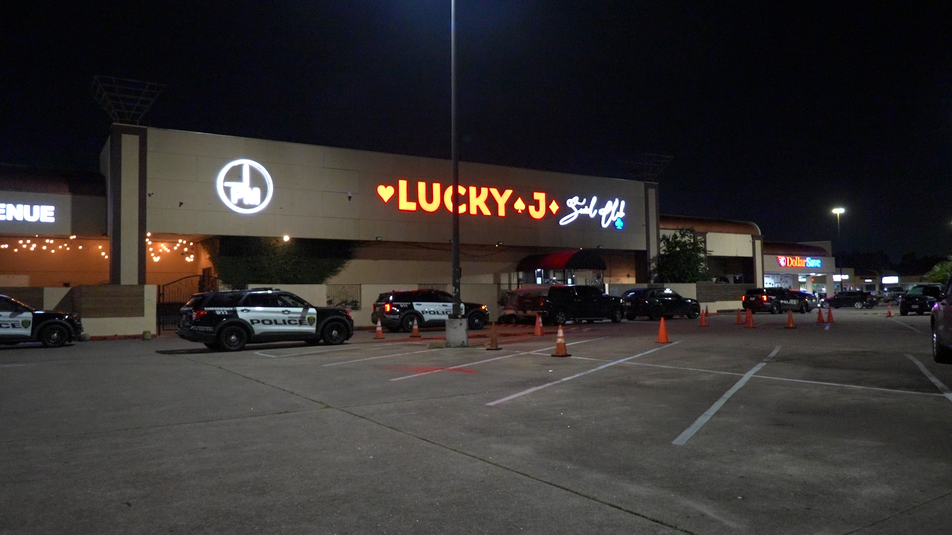 Nighttime parking lot in front of a strip mall with a bright red 'LUCKY' sign, police cars parked along the curb, and orange cones scattered around.