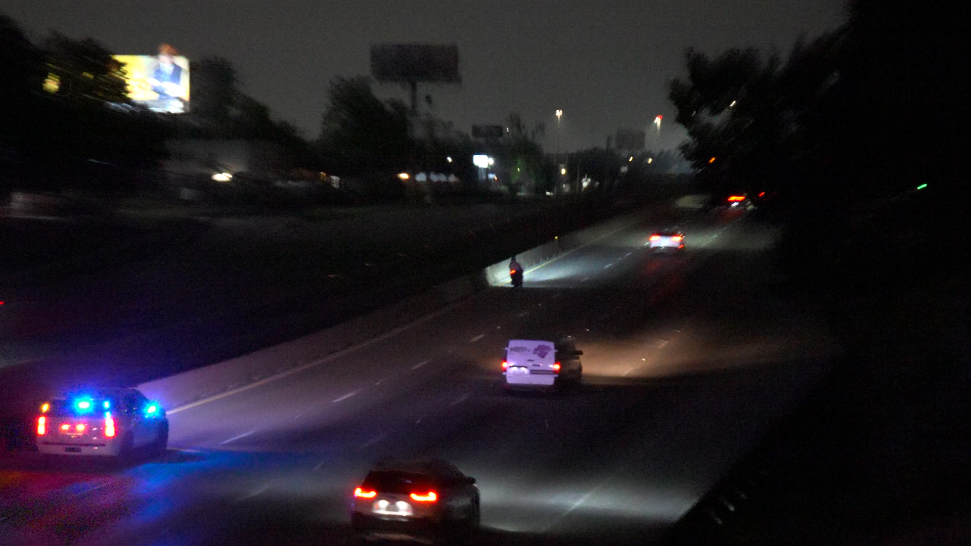 Nighttime freeway scene with a police car flashing blue lights and several cars on the road, including a motorcycle on the shoulder.
