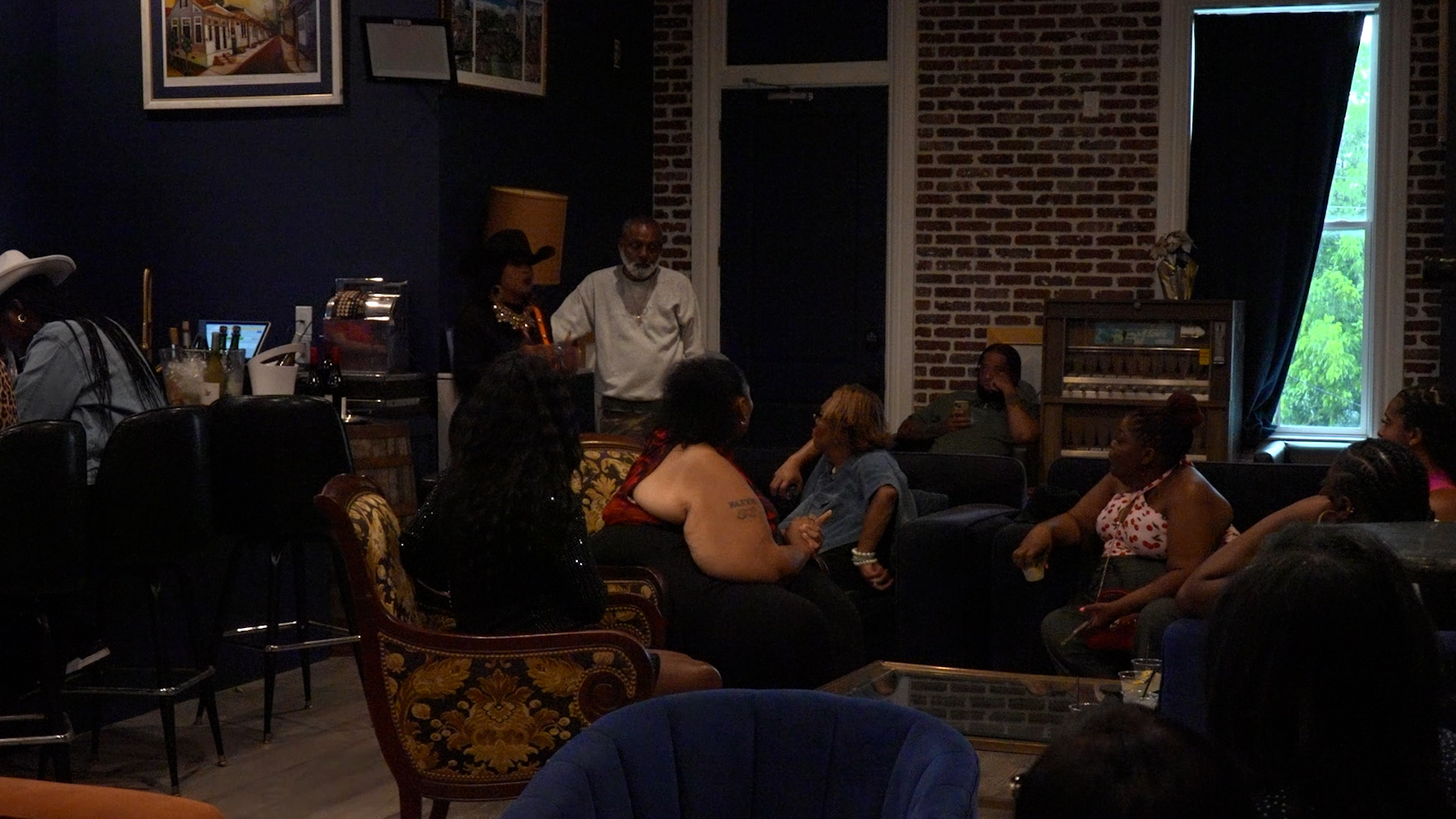 Group of people seated in a dim lounge, watching two individuals talk near a small stage against a brick wall.