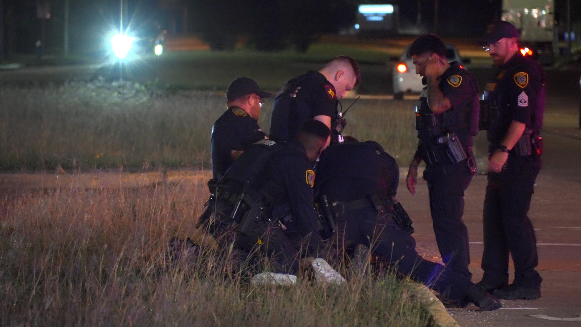 Nighttime scene of several police officers in tactical gear kneeling in a circle on a grassy area, discussing a task.
