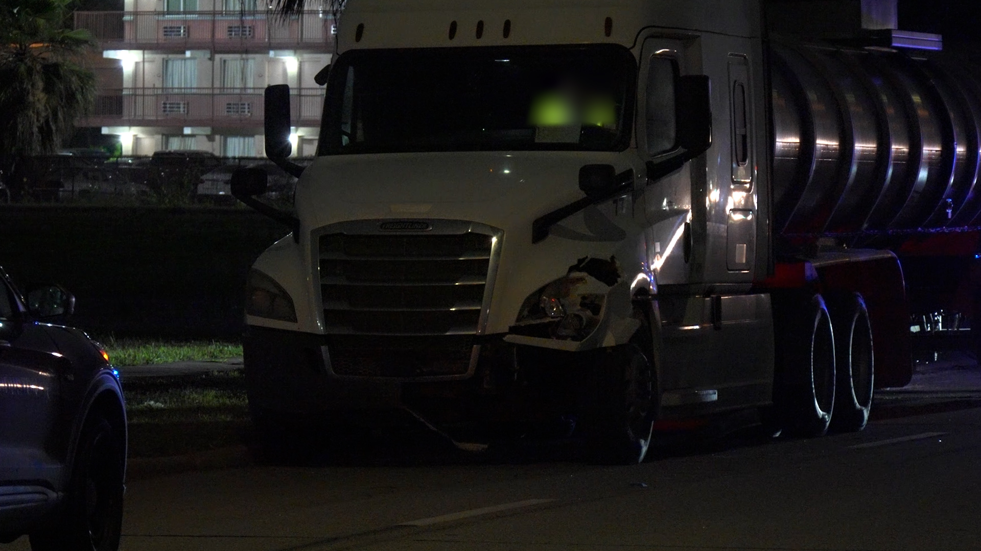 White semi-truck with front-end damage parked at night, tanker trailer attached, emergency lights reflecting nearby vehicles