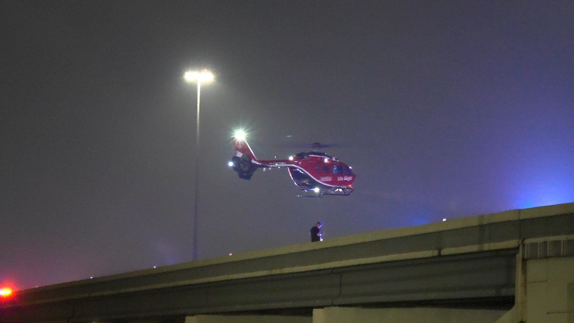 Red Life Flight helicopter hovering above a freeway overpass at night, with blue emergency lights in the distance.