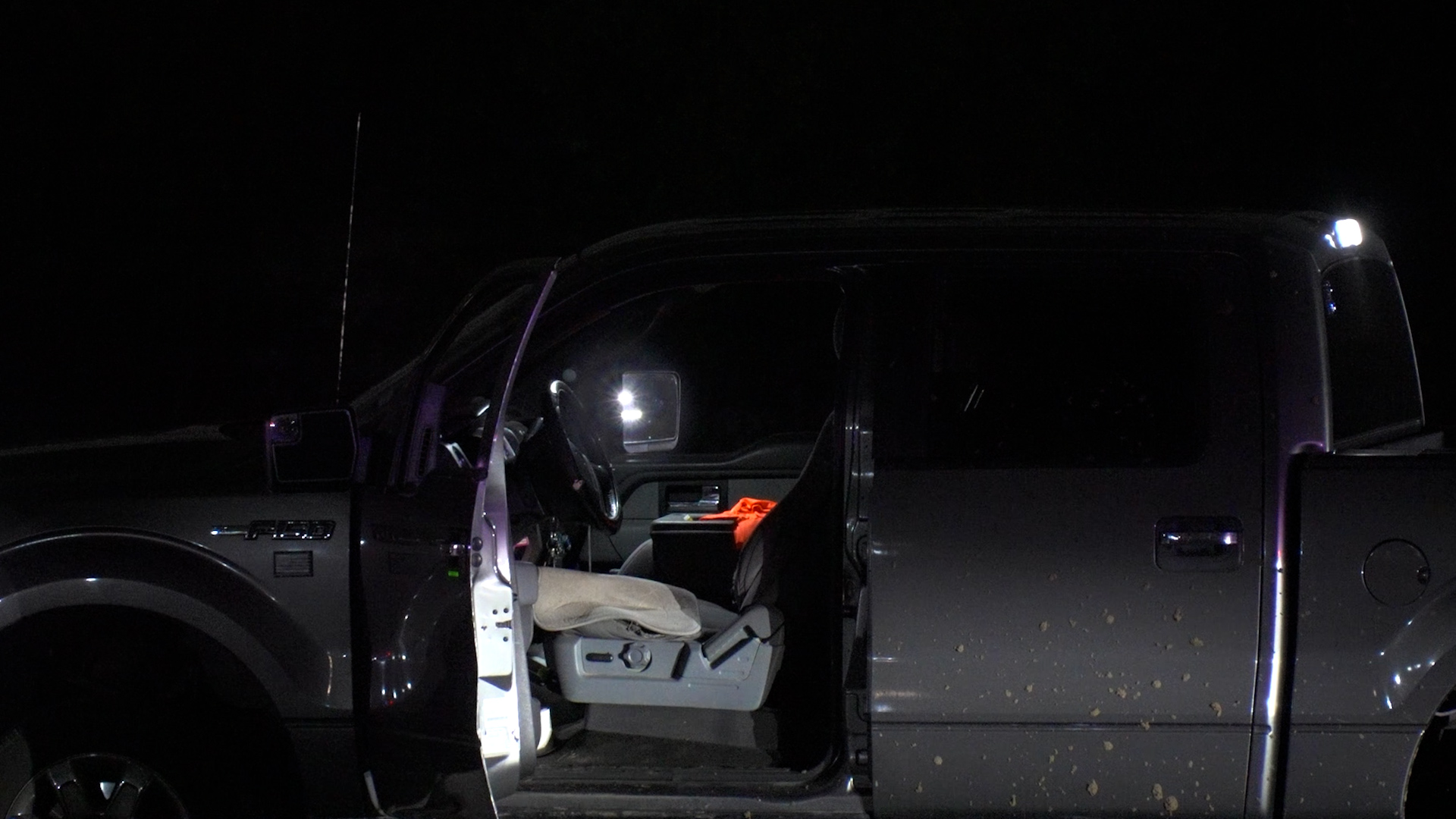 Dark pickup truck at night with the driver’s door open, interior lit by dashboard lights and a bright top-side light visible on the cab.