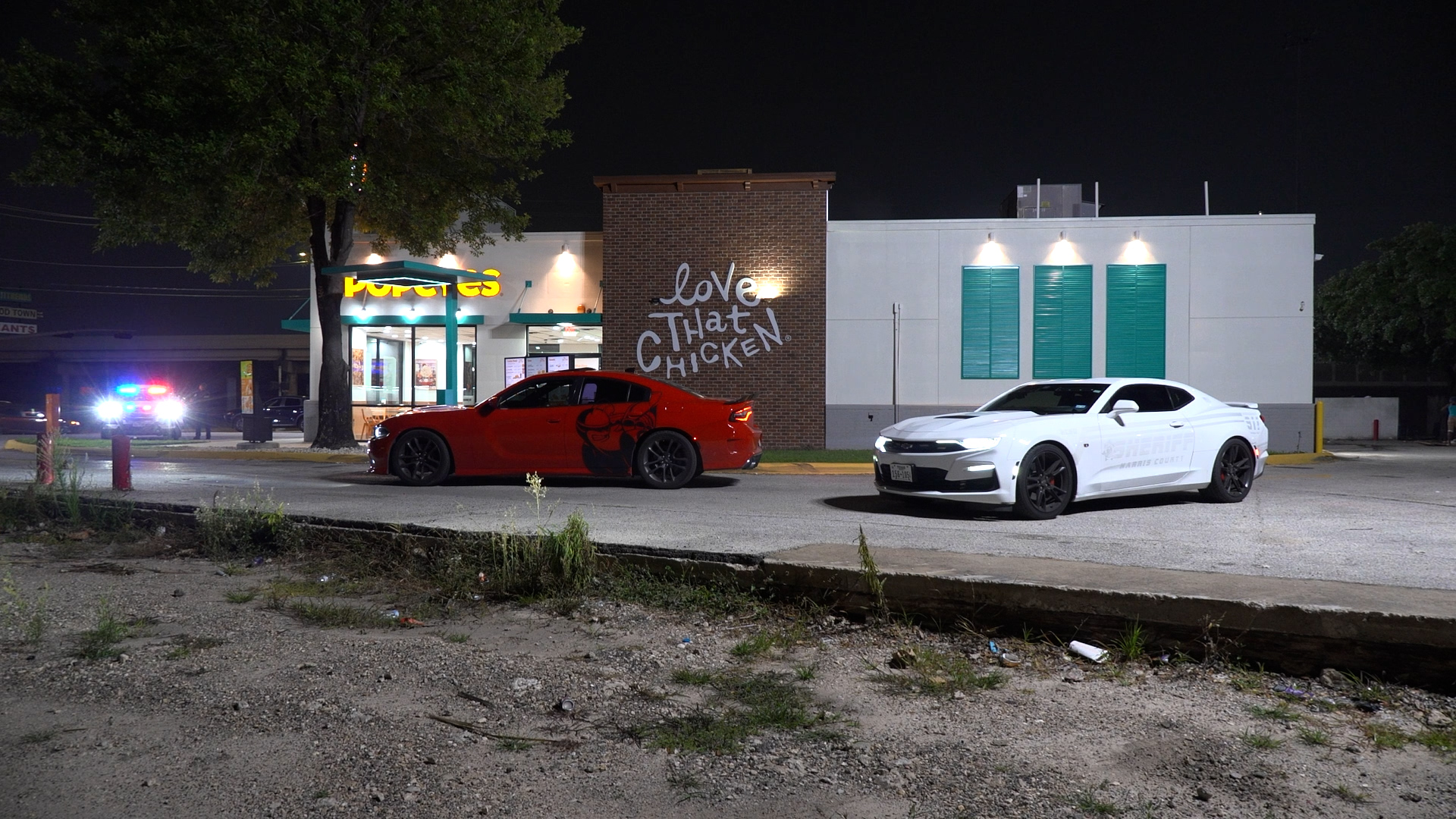 Night scene at a fast-food restaurant with a red sports car and a white sheriff car parked outside; a brick wall mural reads 'Love That Chicken'.