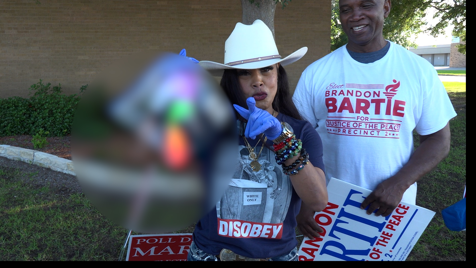 Woman in a white cowboy hat and blue gloves points at the camera during an outdoor protest, with a man in a campaign shirt holding signs nearby and various protest signs visible in the background.