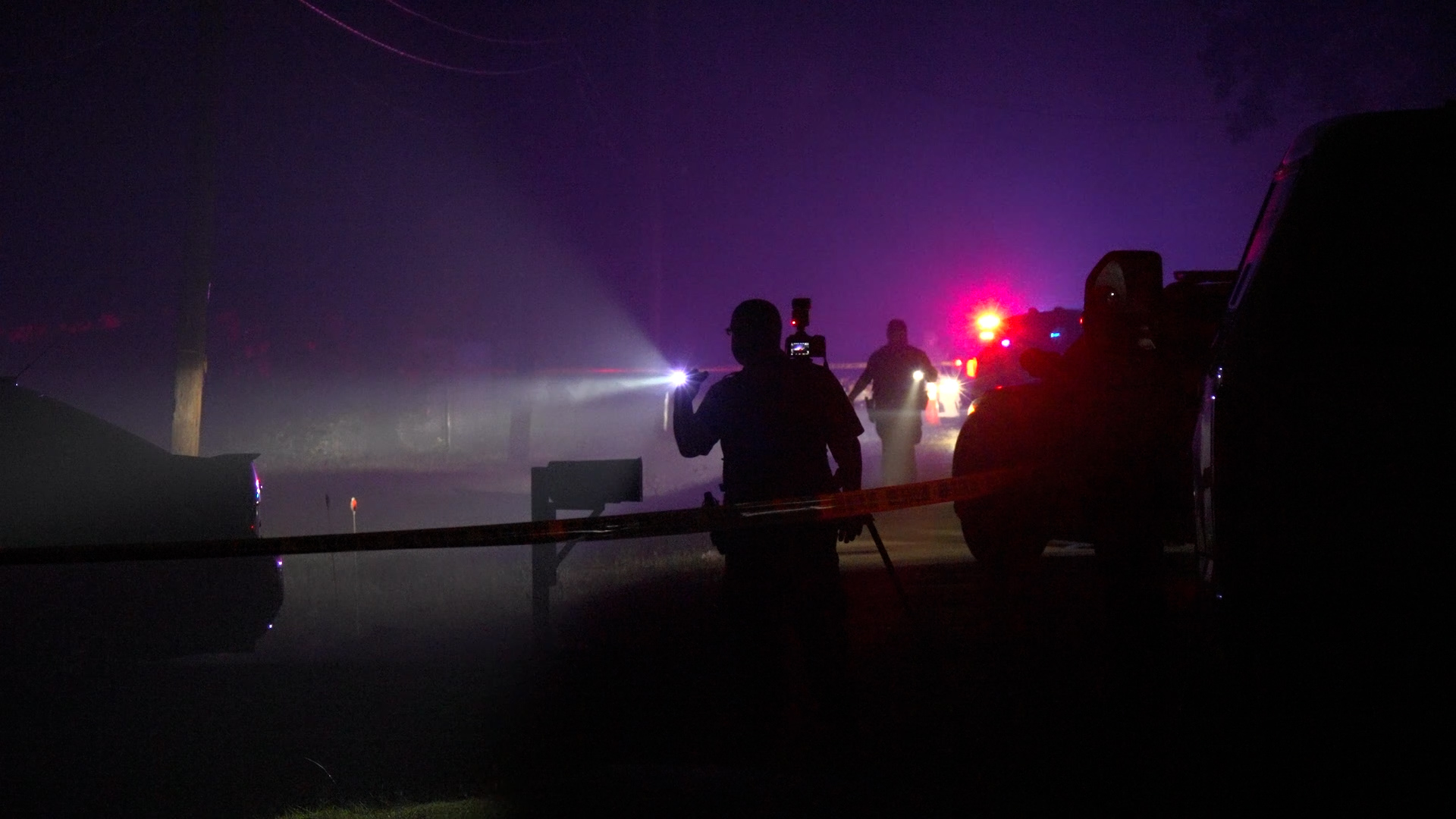 Dark night scene of a police or emergency roadside with flashing red and blue lights, a masked figure holding a flashlight, and yellow tape. Note: concise descriptive alt text.