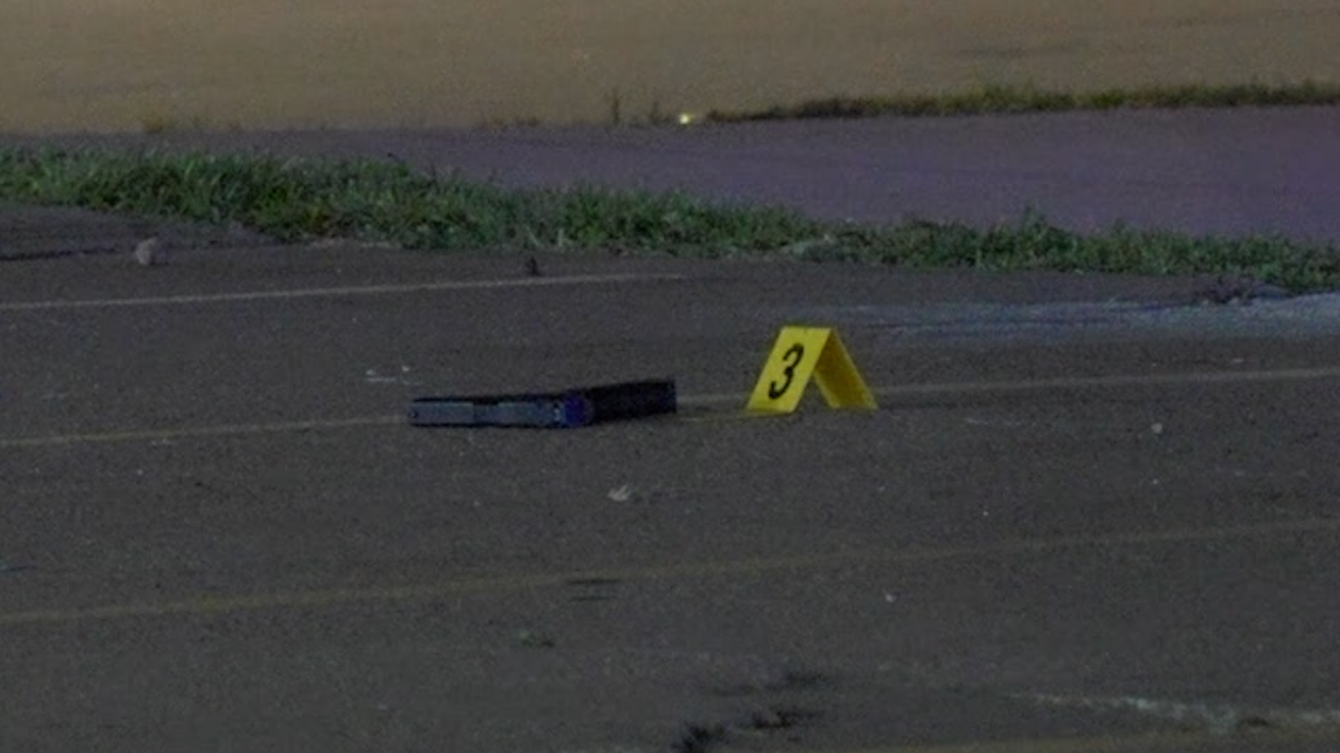 Yellow evidence marker with the number 3 beside a dark rectangular object on a parking lot surface at night/s dusk locale future parking lot edge.