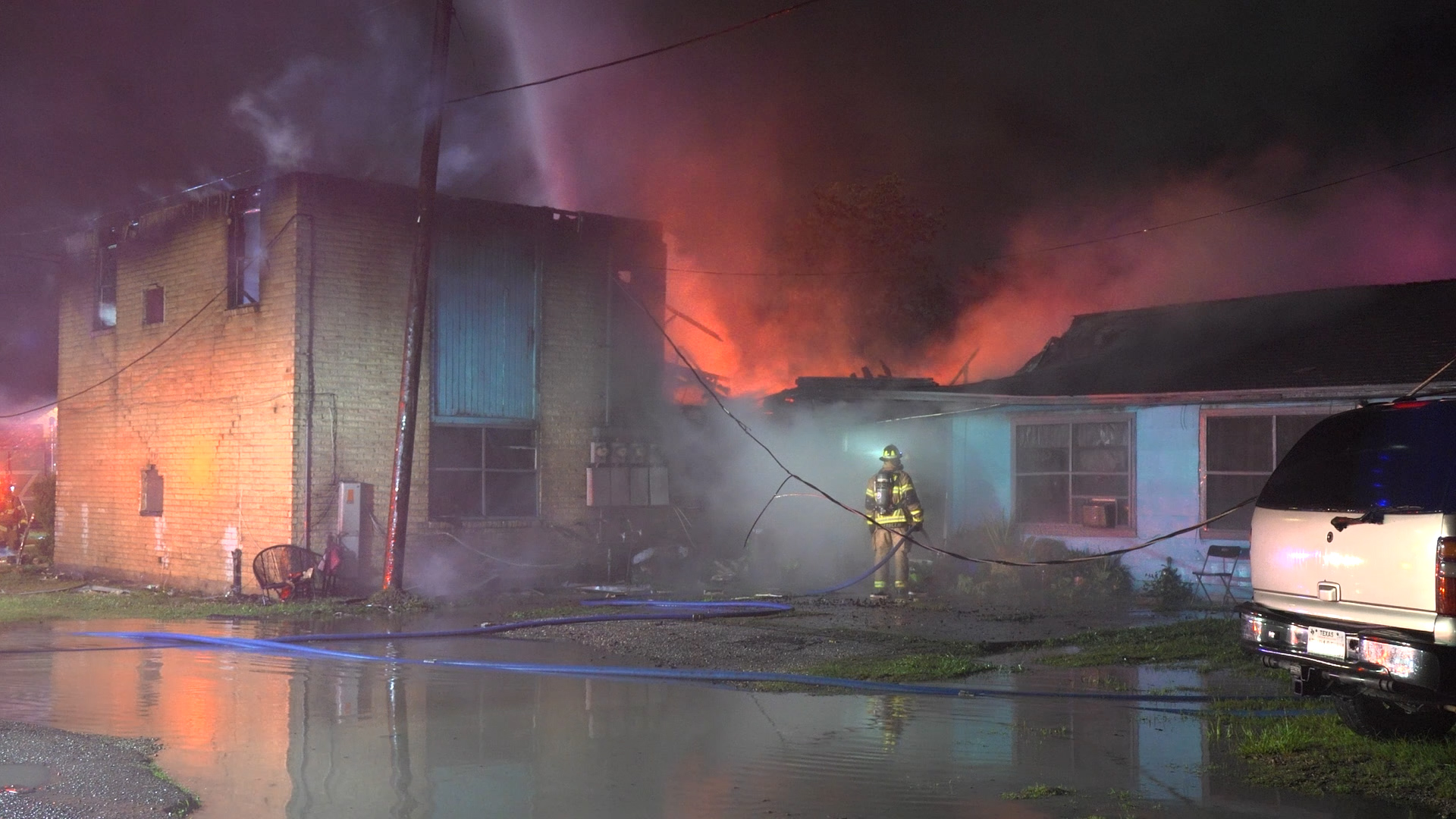Nighttime scene of a brick building on fire with bright orange flames and smoke; a firefighter in gear stands near the blue entrance while water covers the street.