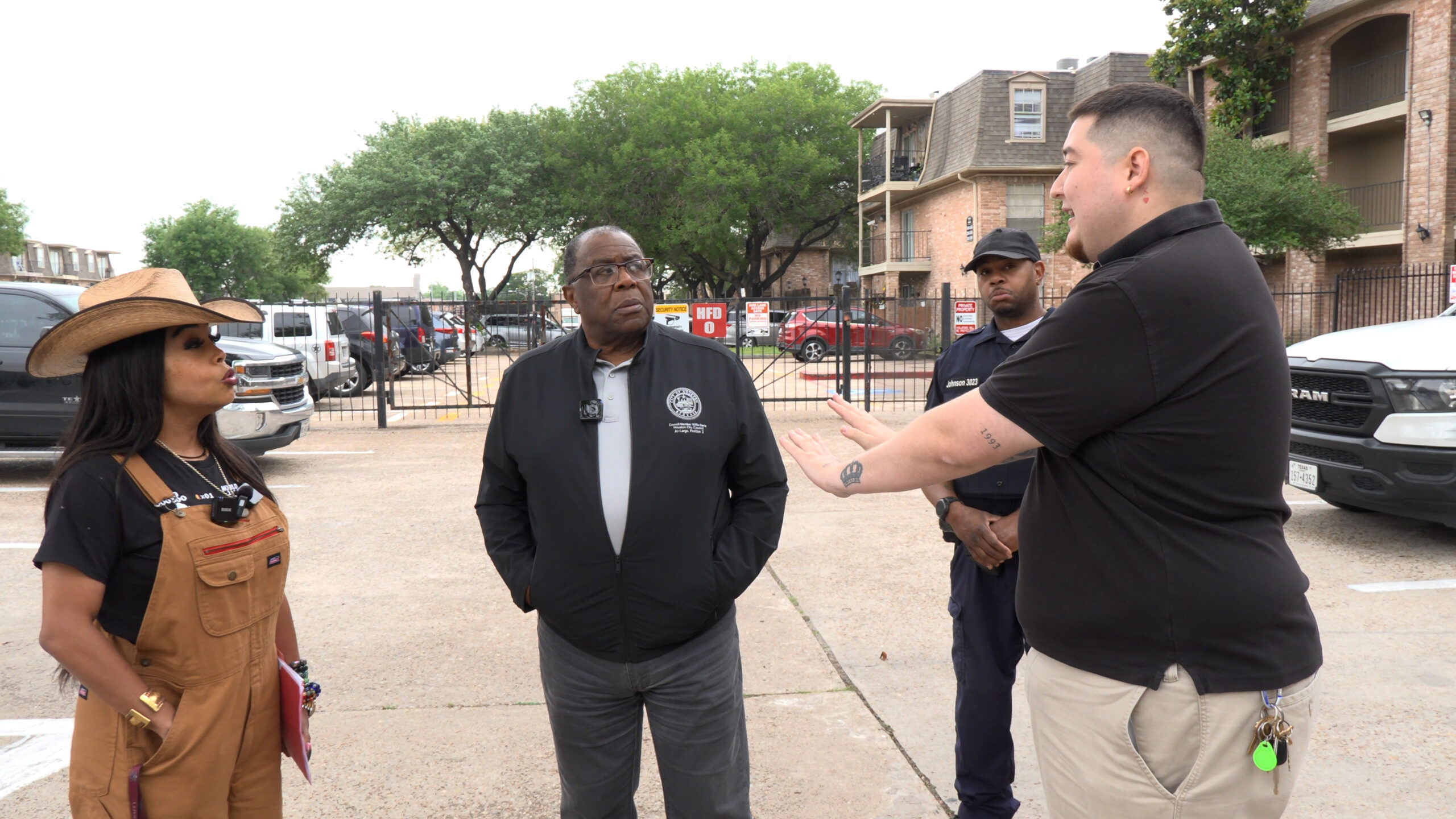 Four people stand in a parking lot having a discussion; a woman wearing a wide-brimmed hat and brown overalls is on the left, and three men listen to her.