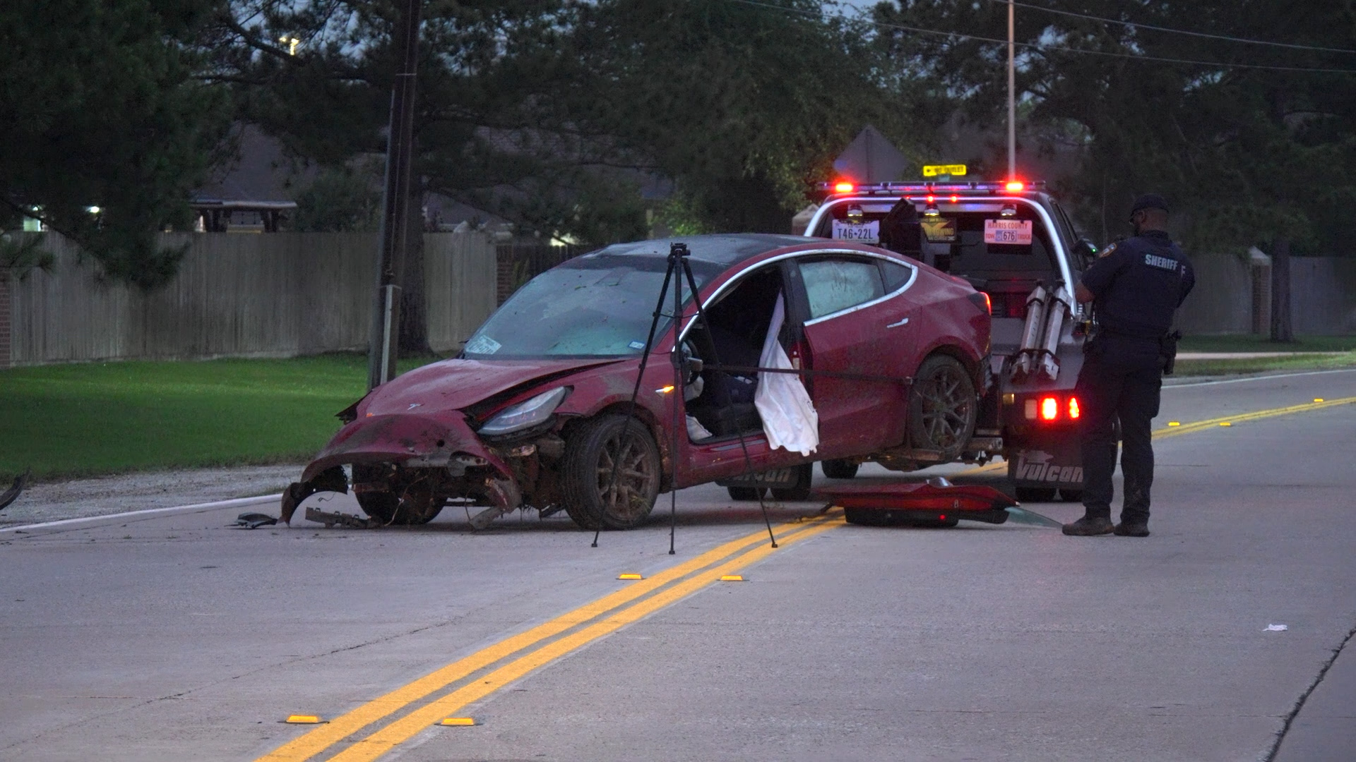 Red sedan heavily damaged in a roadside crash being loaded onto a tow truck; a sheriff's deputy stands nearby with red flashing lights in the background.