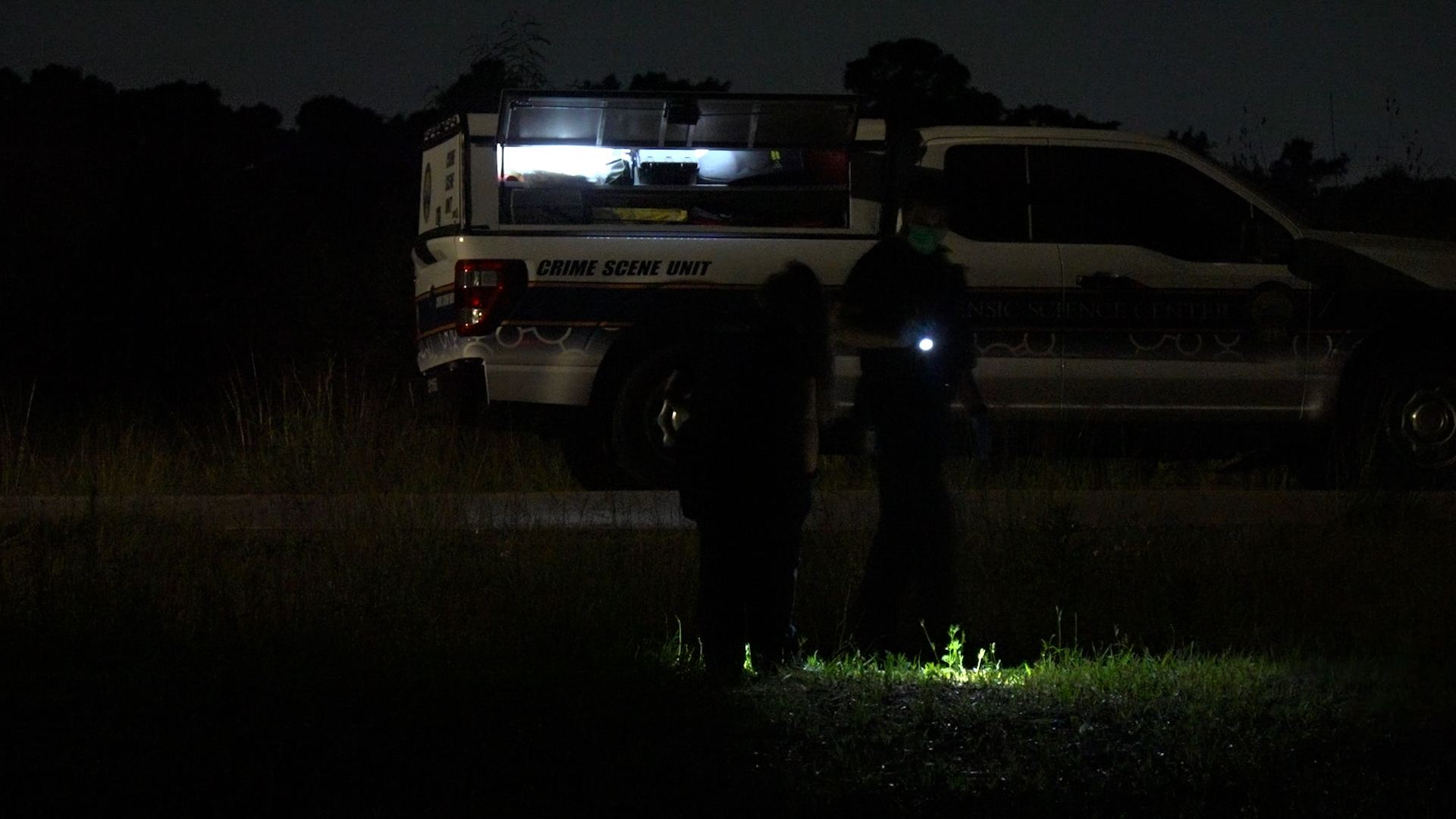 Crime Scene Unit van at night with its back hatch open and bright interior lights, two investigators silhouetted nearby with a flashlight.