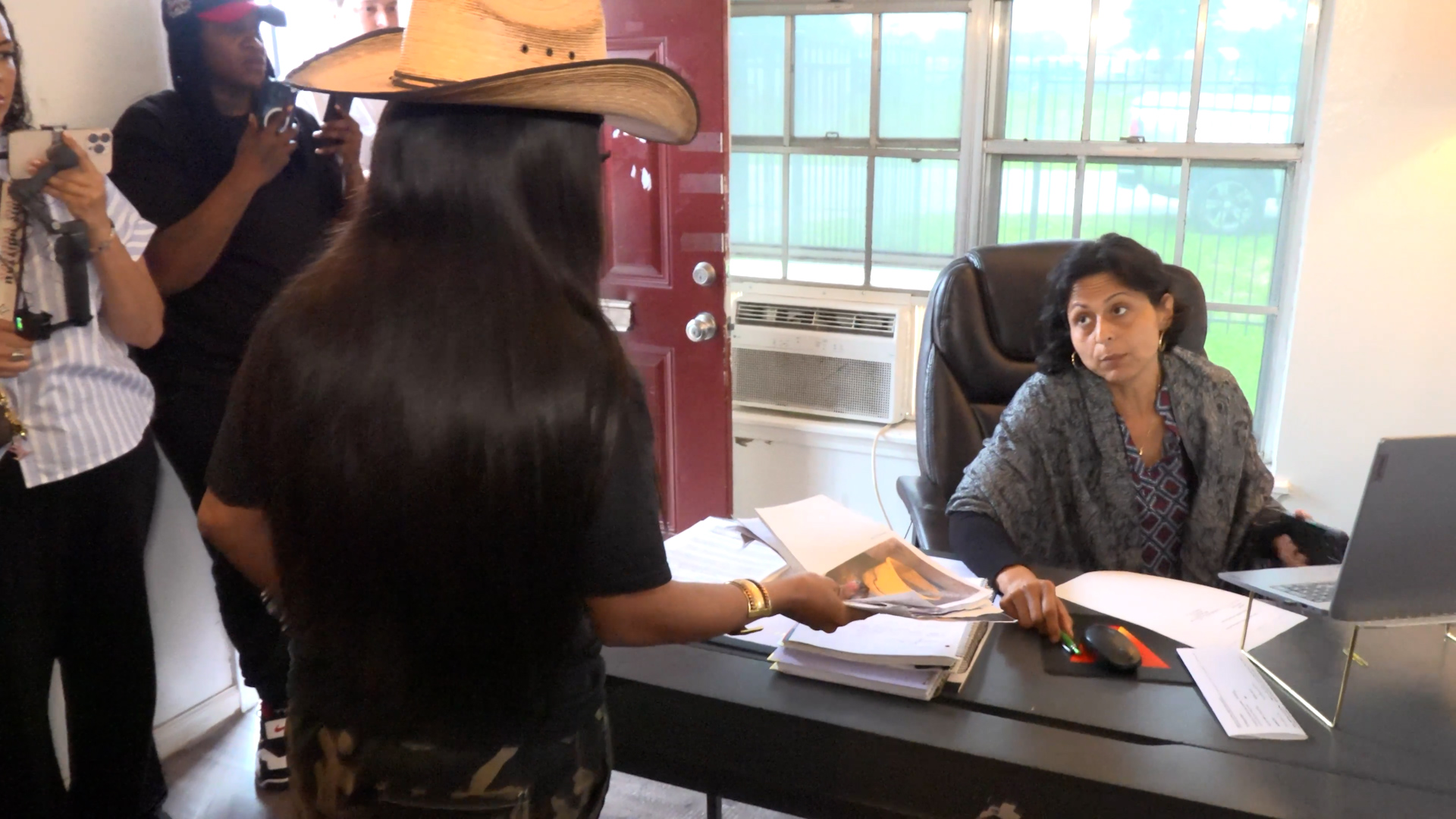 Office scene: woman with dark hair seated at a desk with laptop and papers, while a person wearing a wide-brimmed hat stands nearby holding documents.