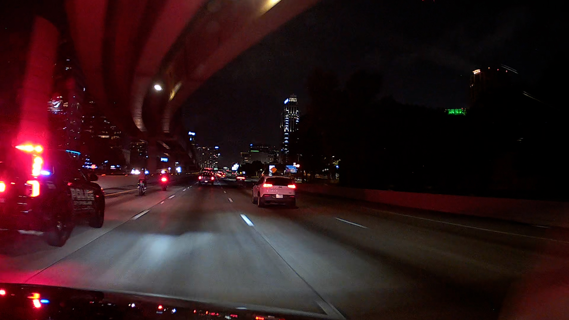 Nighttime urban highway with red emergency lights on left and city skyline in distance.