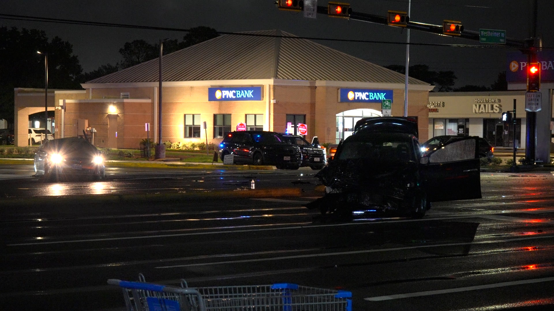 Nighttime crash scene in a bank parking lot; damaged SUV in the foreground, police cars with flashing lights by a PNC Bank.