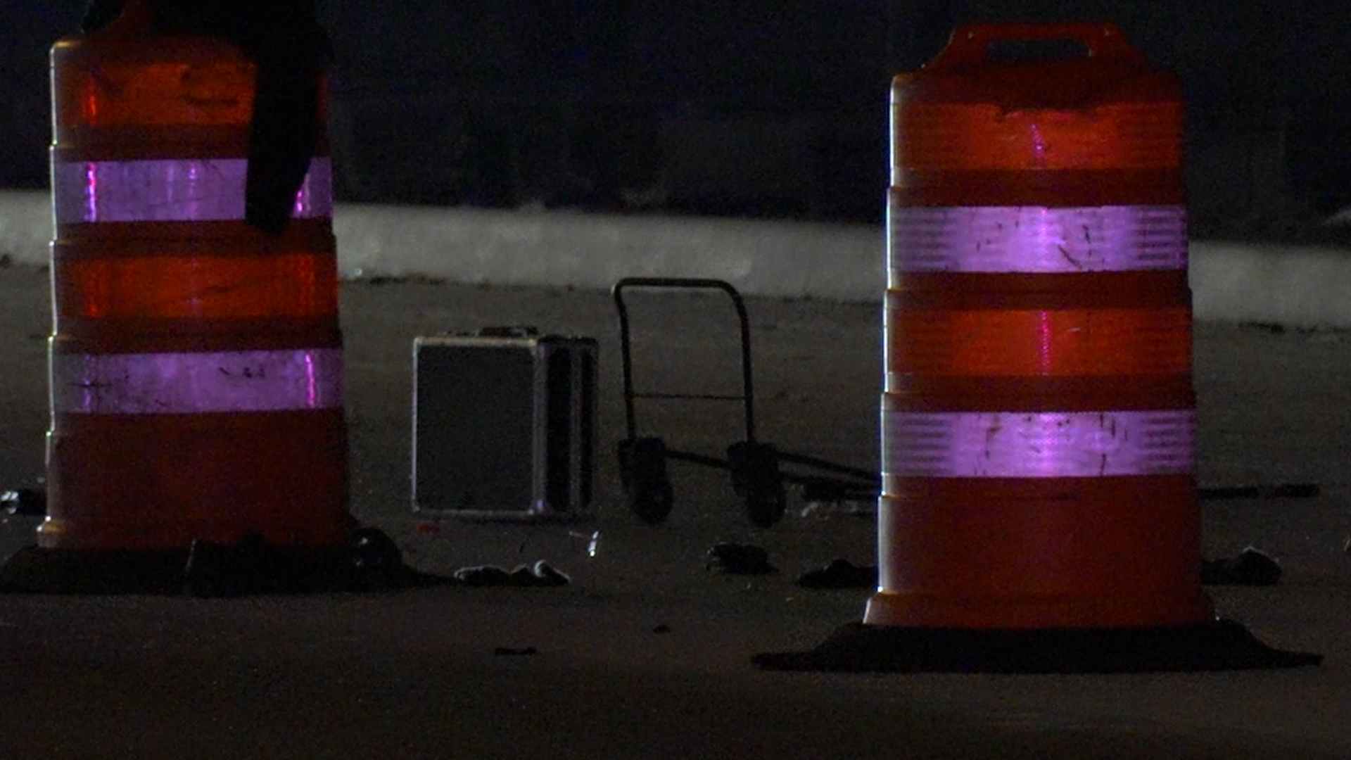 Nighttime roadside scene with orange and white construction barrels and a hand truck in the background.