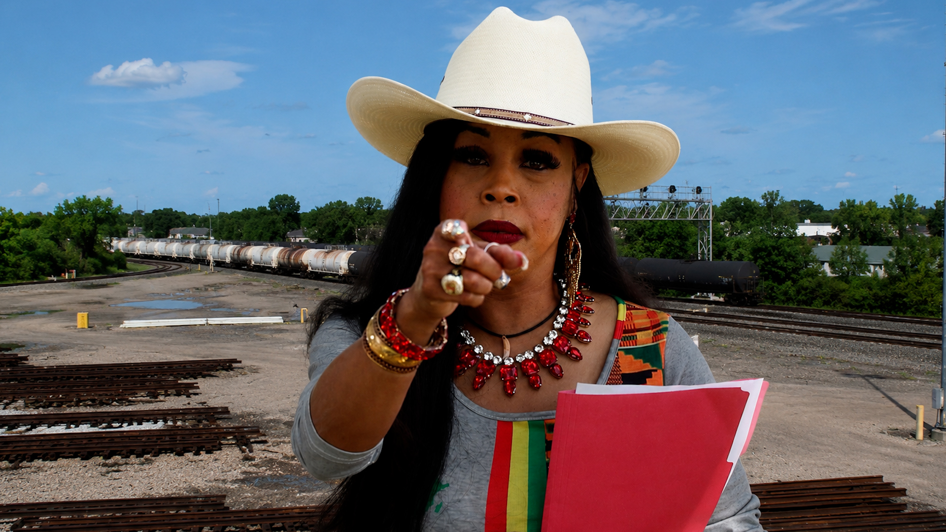 Woman wearing a large white hat and red lipstick points at the camera in a rail yard with stacked train cars in the background, holding pink folders/canvases in her arms.