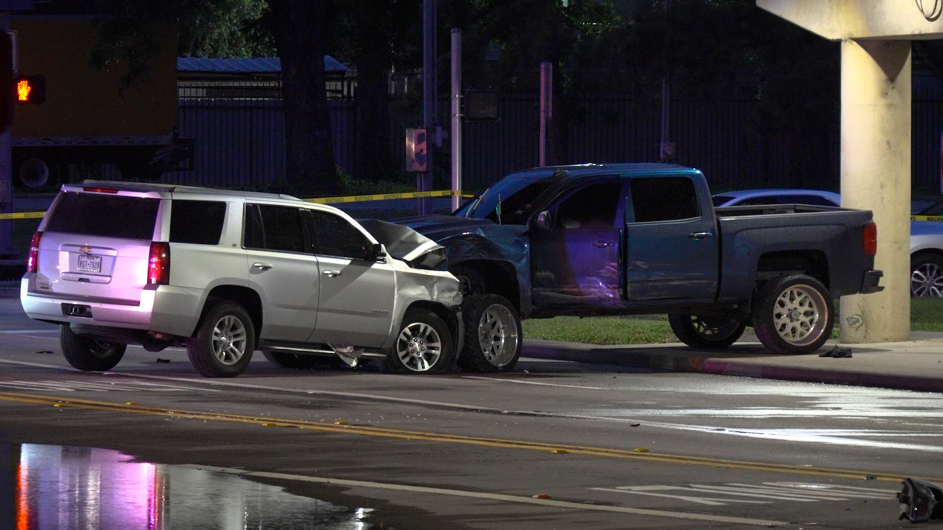 Nighttime crash scene with a white SUV and a blue pickup truck, front-end damage, caution tape, and wet reflections on the street.