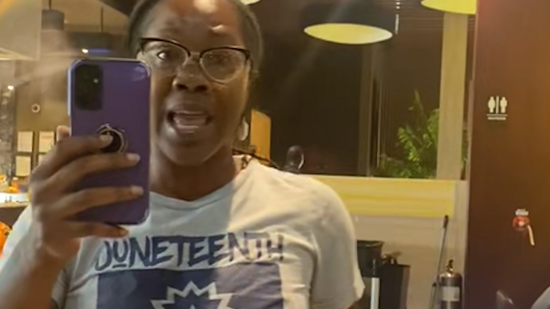 Person wearing glasses takes a mirror selfie with a purple smartphone in a cafe or restaurant setting