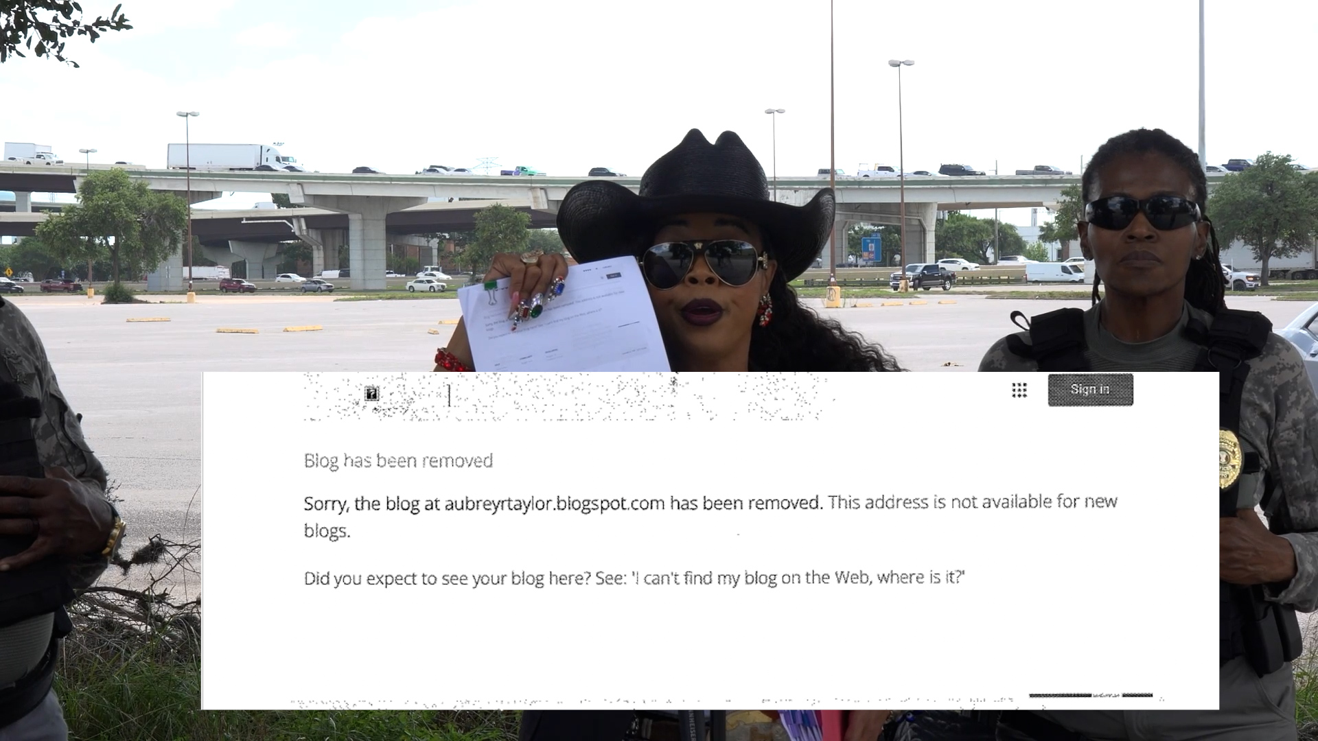 Two women in sunglasses stand in a parking lot by a highway overpass; the left woman wears a black cowboy hat and holds papers.