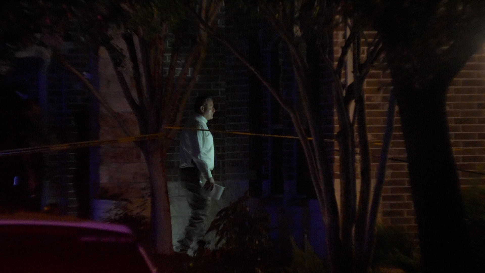 Nighttime scene with a man in a light-colored shirt and pants standing near a brick building, behind yellow caution tape, dimly lit by streetlight.