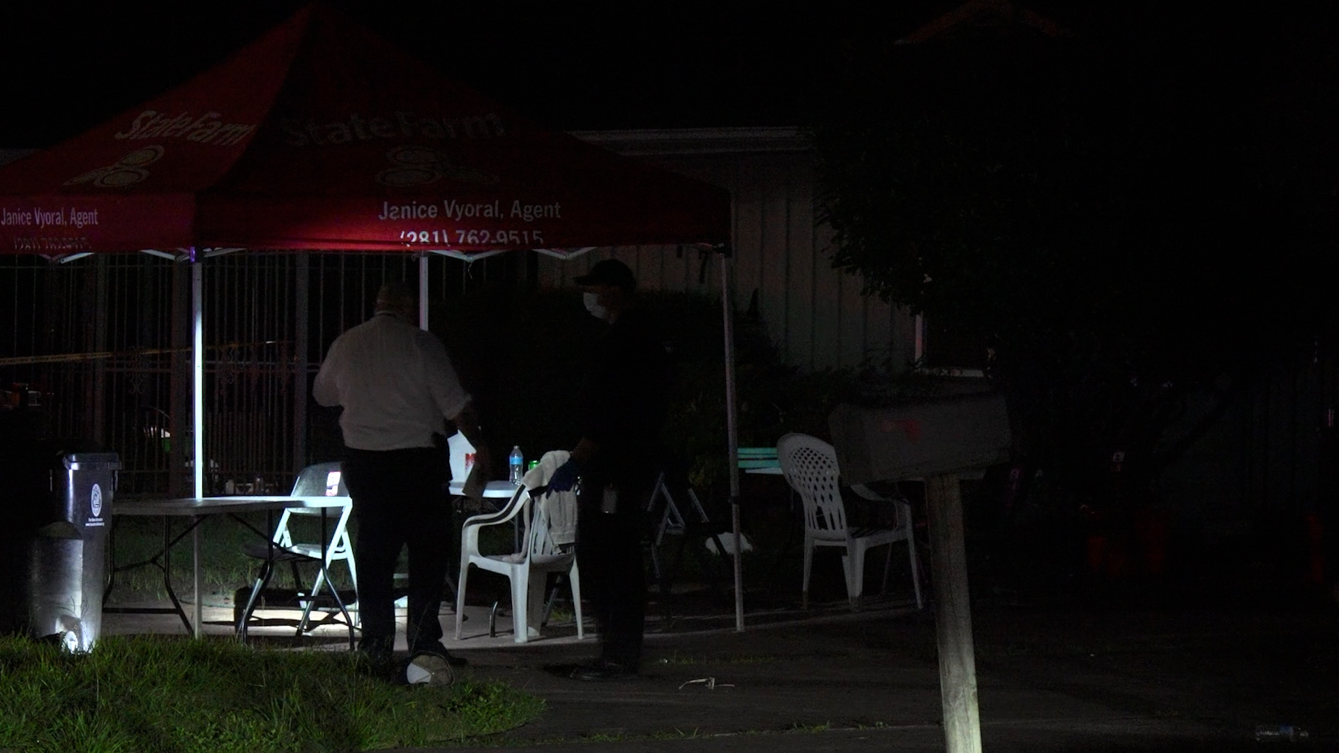 Nighttime outdoor scene under a red canopy with two people standing near a table and plastic chairs; one person wears a mask.