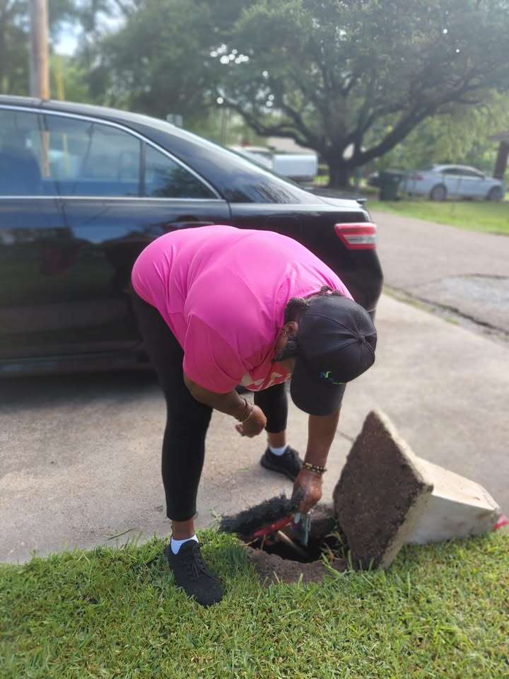 Person in a pink shirt bends over an open manhole in a residential street, lifting a heavy metal cover near a parked car.