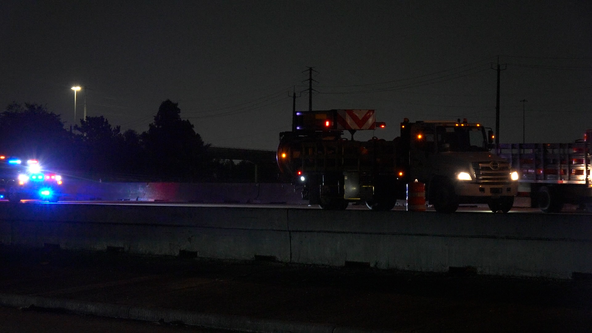 Night highway scene with a large maintenance/utility truck and orange traffic barrels, illuminated by blue and red emergency lights at dusk.