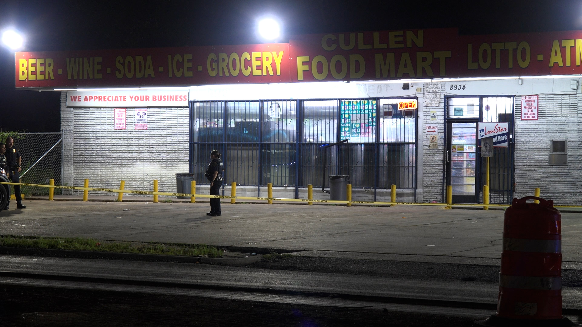 Nighttime exterior of a convenience store with yellow crime tape and police officers guarding the scene outside the storefront windows.
