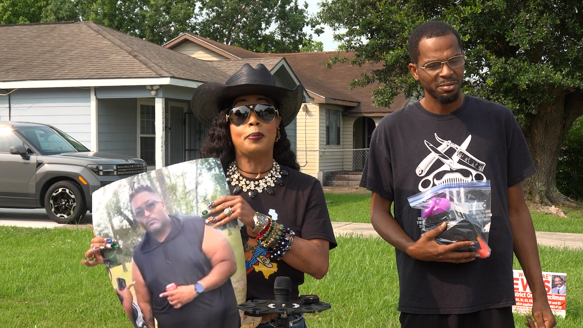 Woman wearing a black hat and sunglasses holds up a photo collage while a man in a dark T-shirt stands beside her, holding a plastic bag of items in a suburban yard.
