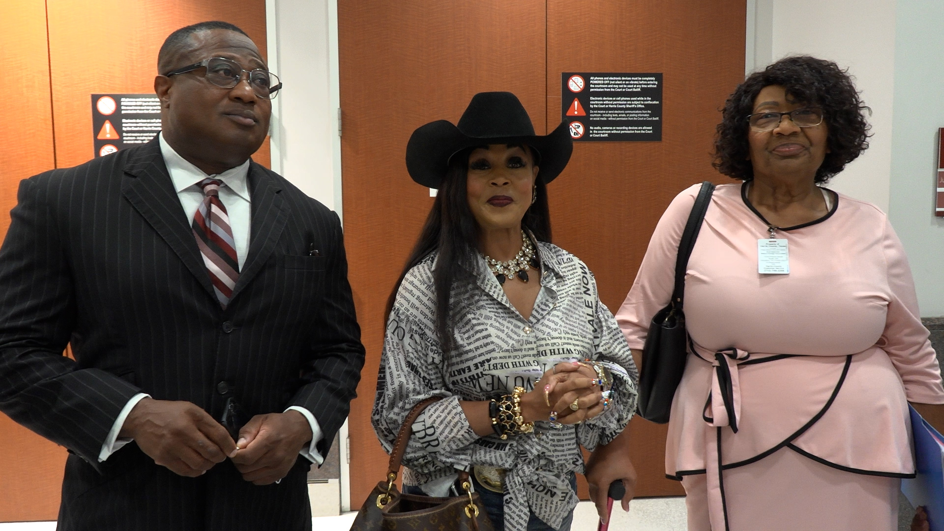 Three adults standing together in a hallway or lobby: a man in a dark pinstripe suit and striped tie on the left, a woman wearing a black hat and patterned shirt in the center, and another woman in a light pink dress on the right with a badge.