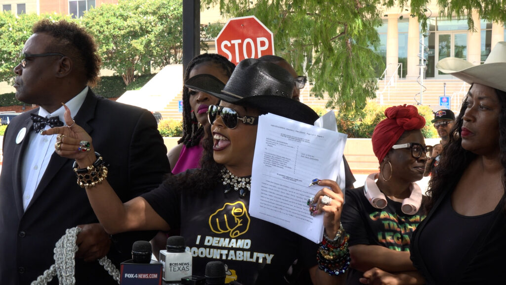 Group of protesters and reporters outdoors; woman in a black hat speaks into microphones, holding papers, with a stop sign behind.