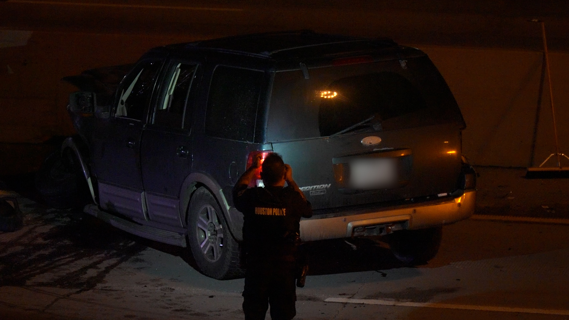 Black SUV crashed on a dimly lit street; a Houston Police officer in the foreground is taking photos of the scene.