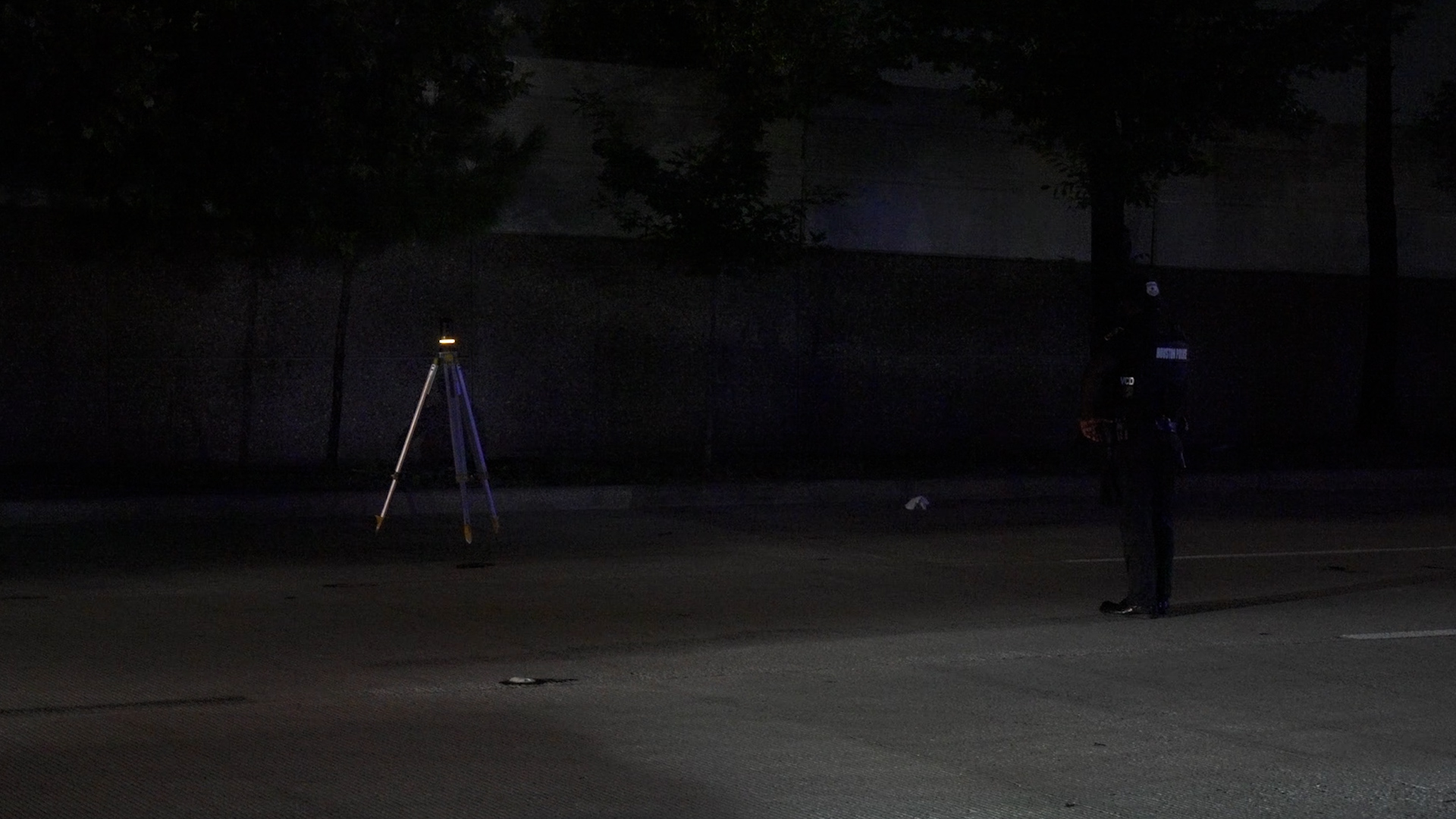 Dimly lit night scene in a parking lot: a tripod with a small light on the left and a uniformed officer standing at right center, silhouette visible.