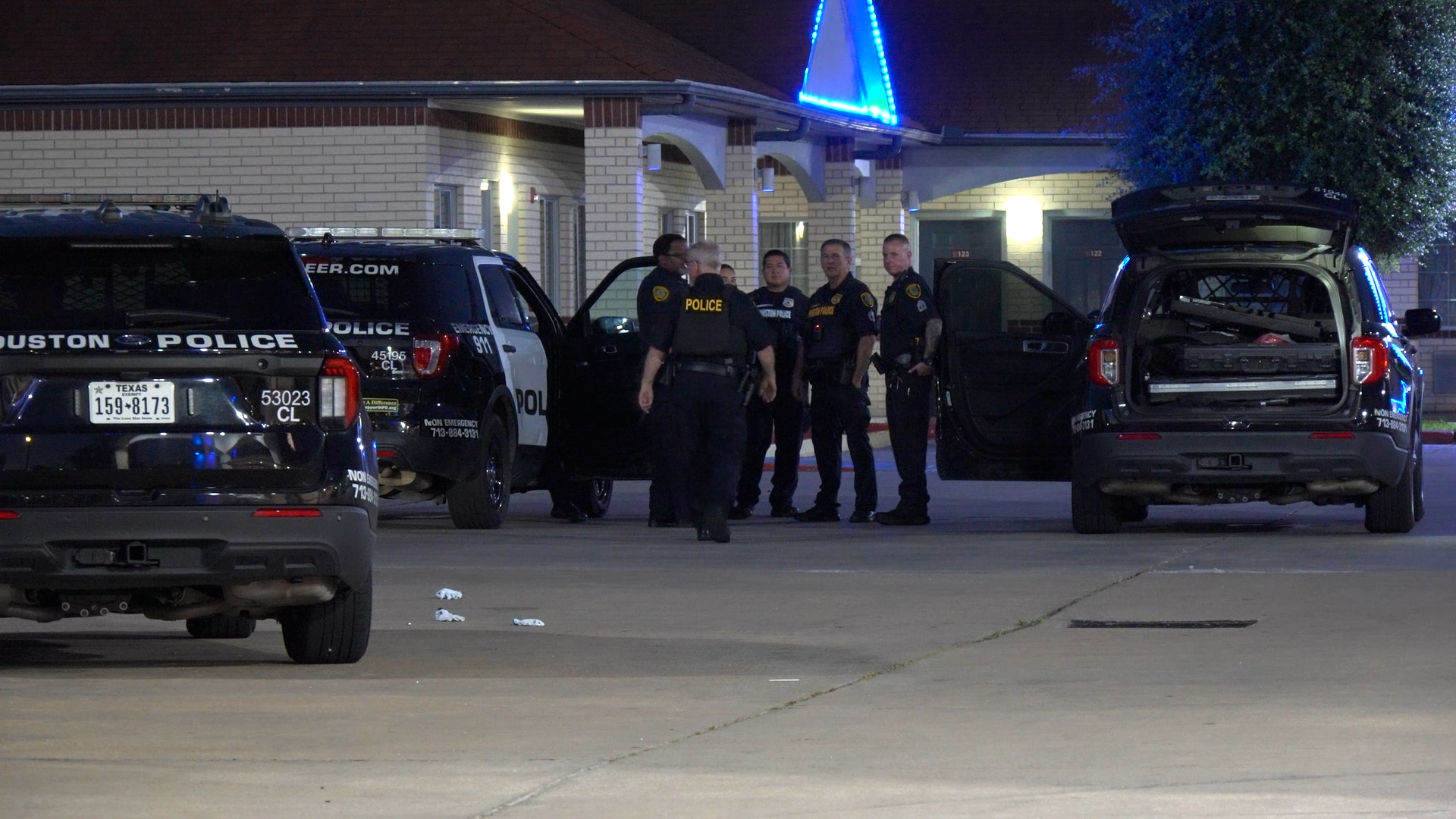 Night scene outside a police station with several Houston Police vehicles and officers gathered near open car doors under blue lights.