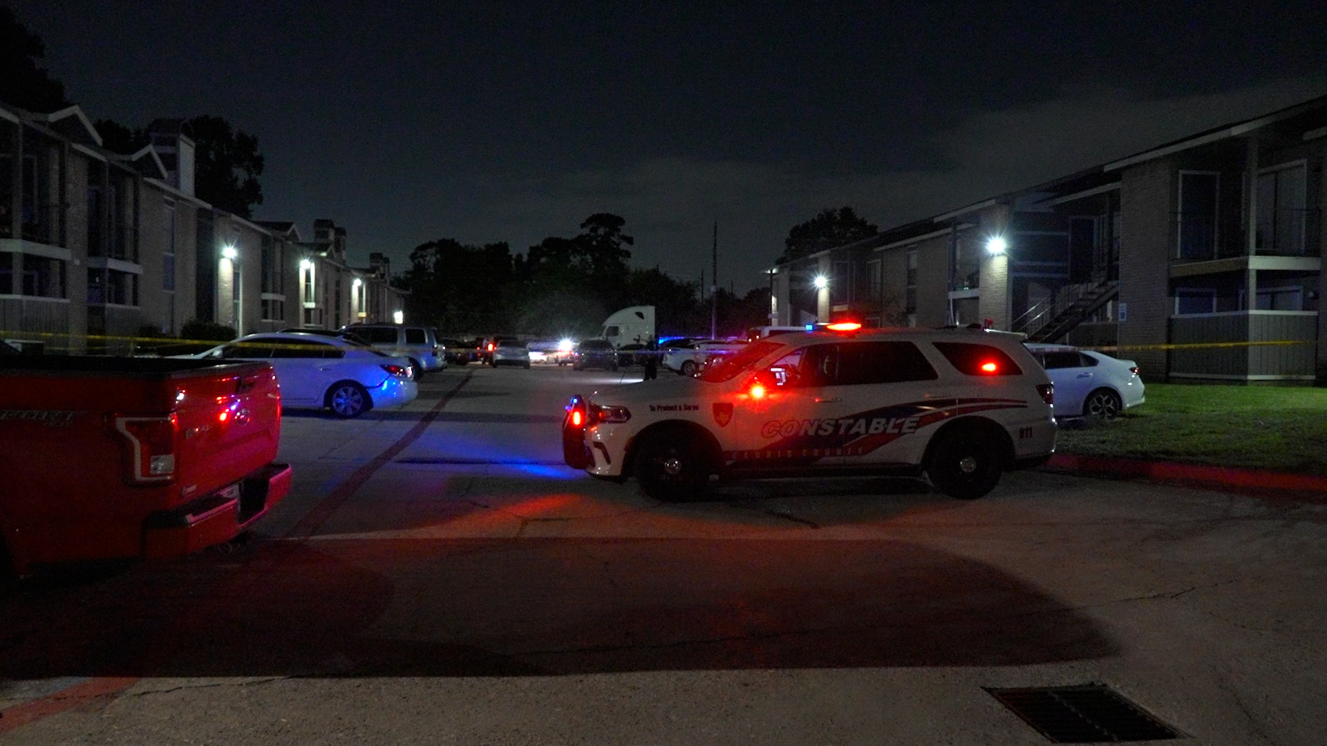 Nighttime residential parking lot with police SUV and flashing red/blue lights, yellow tape marking off an area, row of apartment buildings in the background.