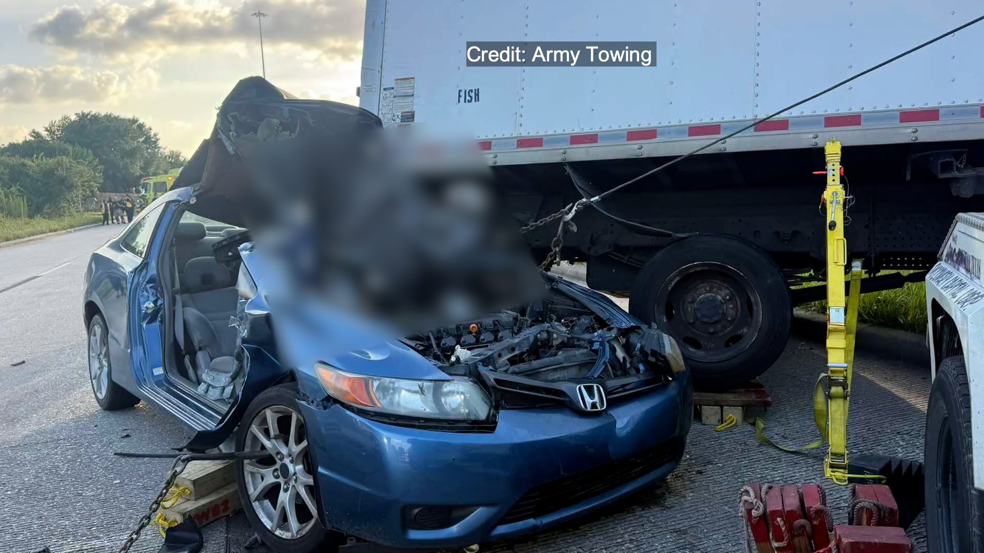 Blue Honda with severe front-end damage sits next to a large white trailer, secured for towing on a highway scene