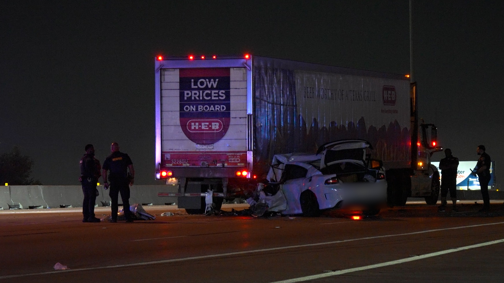 Nighttime highway crash scene with a crushed white car under a large trailer, police and responders on scene.