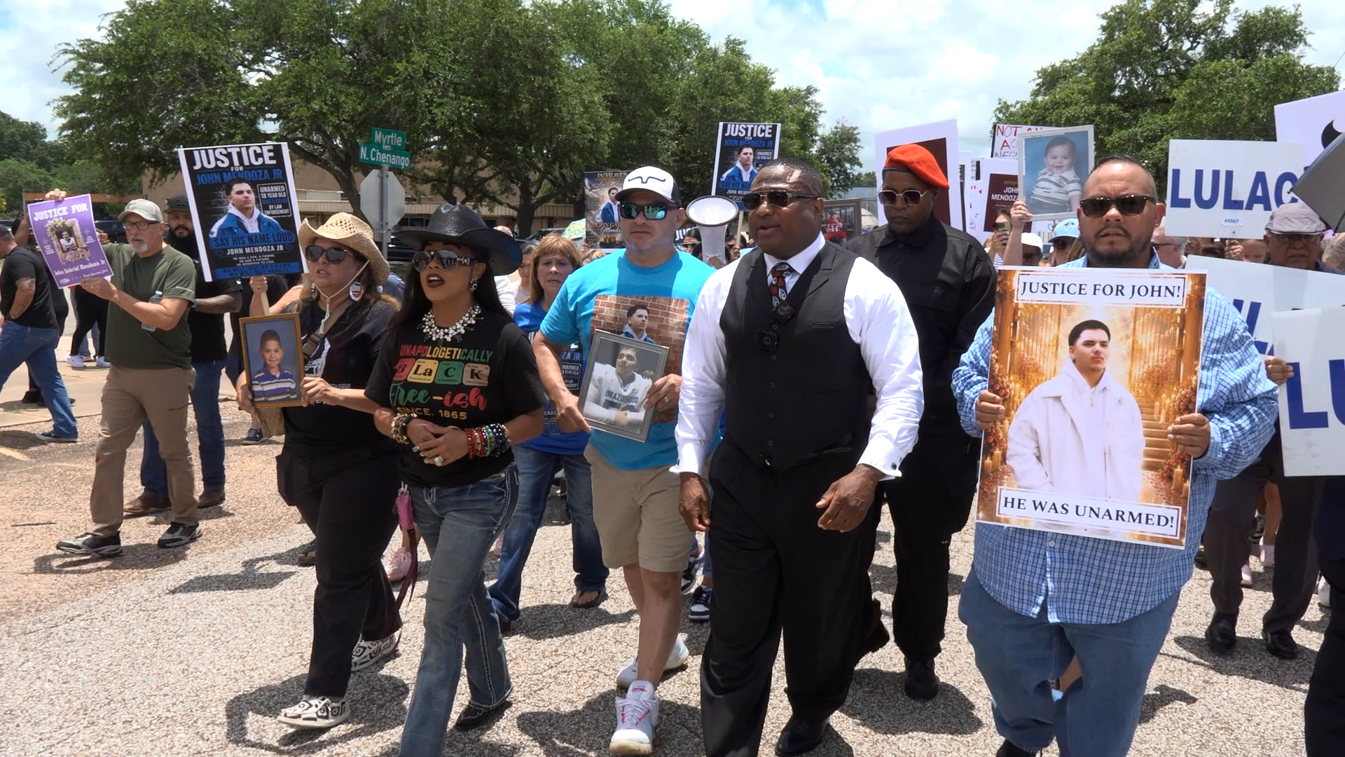 Group of protesters marching on a sunny street, holding posters and photos demanding justice for John Mendoza Jr.