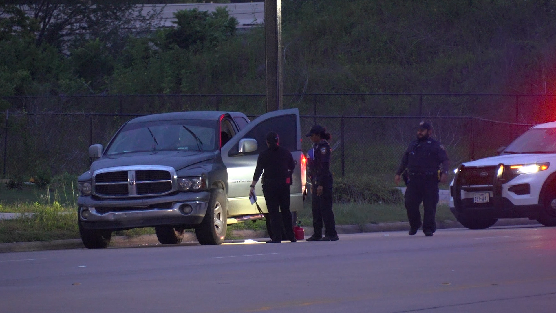 Police officers examine a gray pickup with its driver’s door open as a patrol SUV with red lights glows in the background at dusk/early evening.