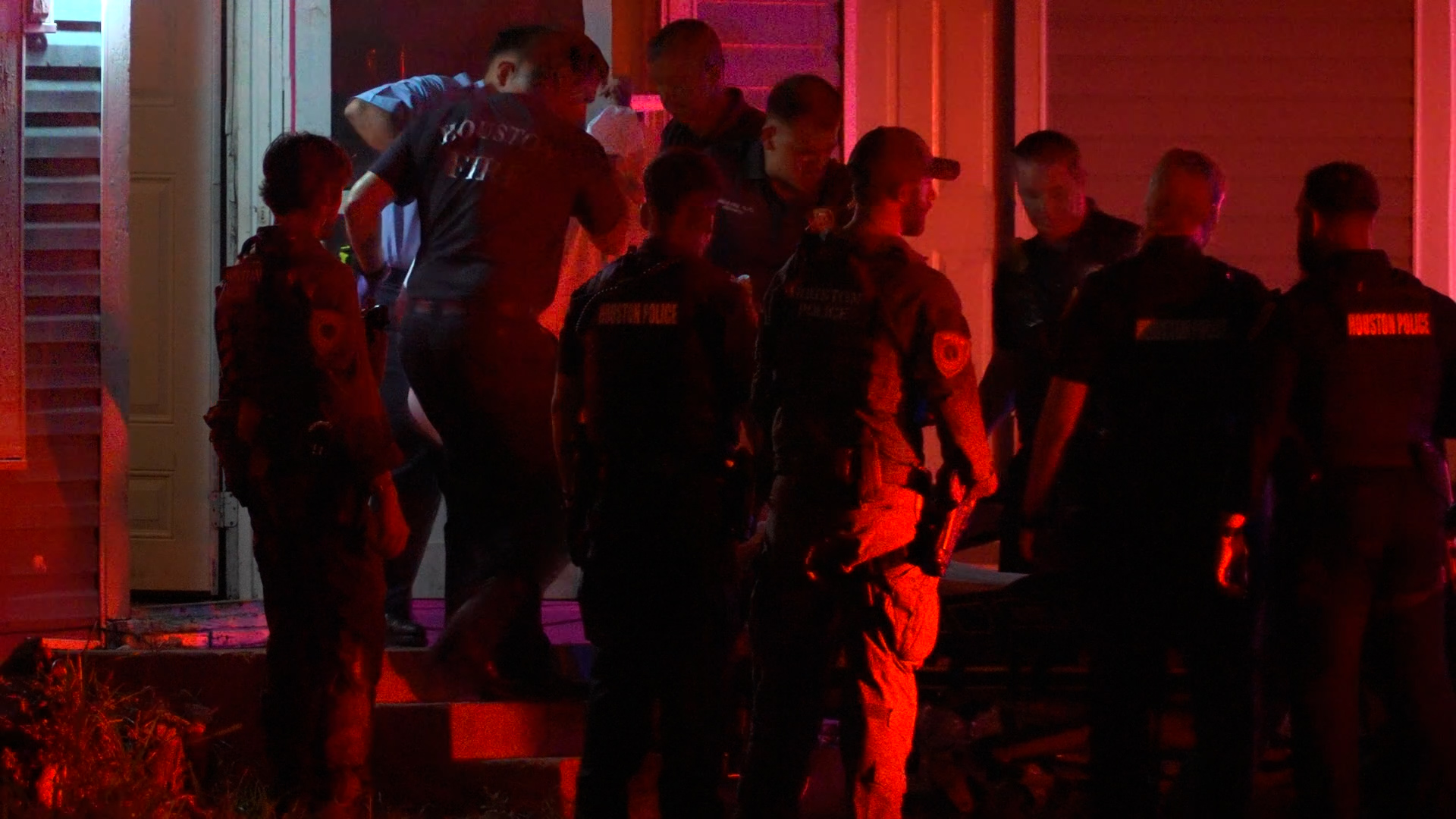Police officers in tactical gear gather outside a doorway at night under red and blue emergency lights.