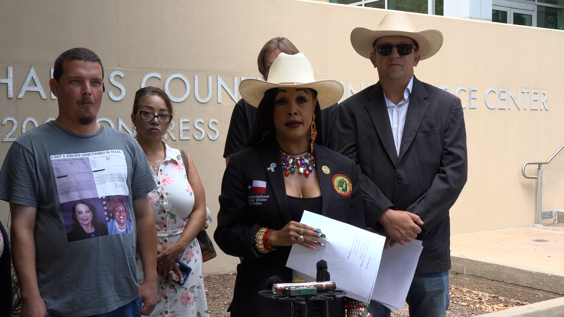 Group of five people standing outside a beige wall; woman in a white hat and bold jewelry speaks to a small press with microphones in front of her.
