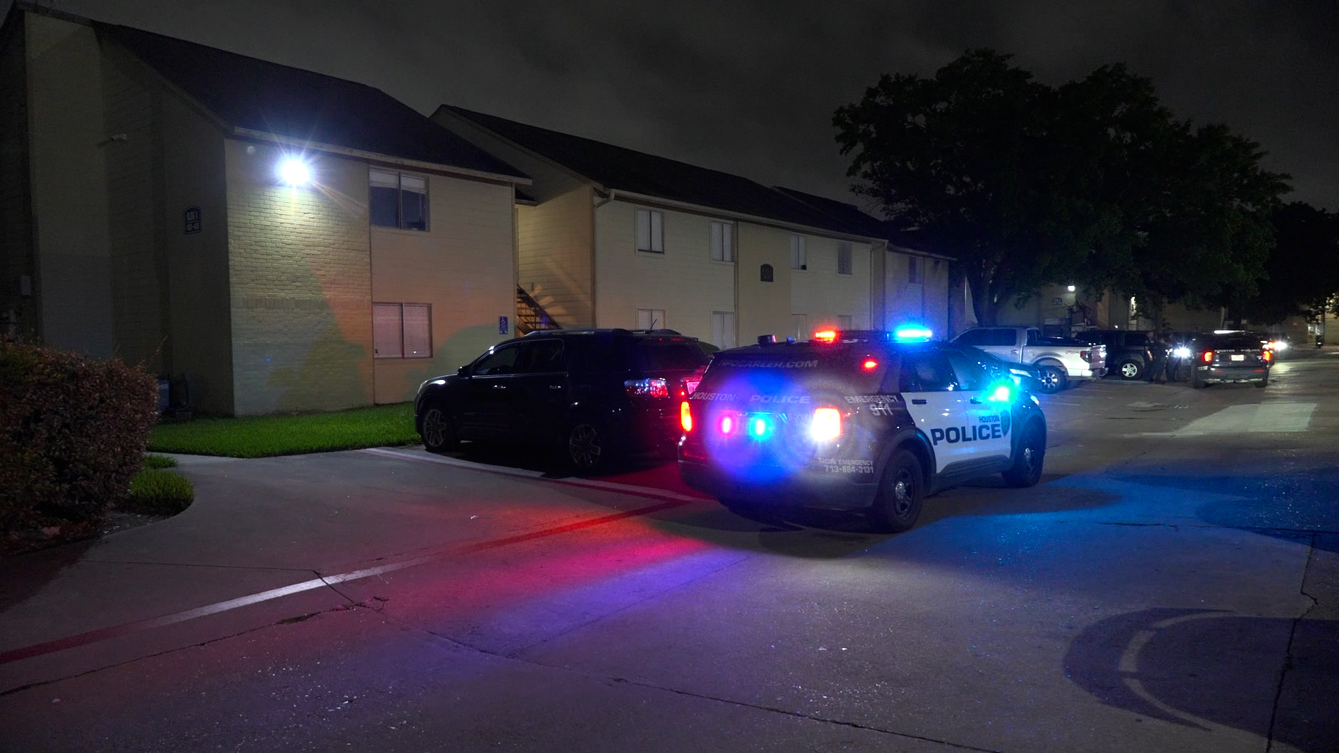 Nighttime scene of a Houston Police SUV with red and blue emergency lights in a parking lot beside an apartment building.