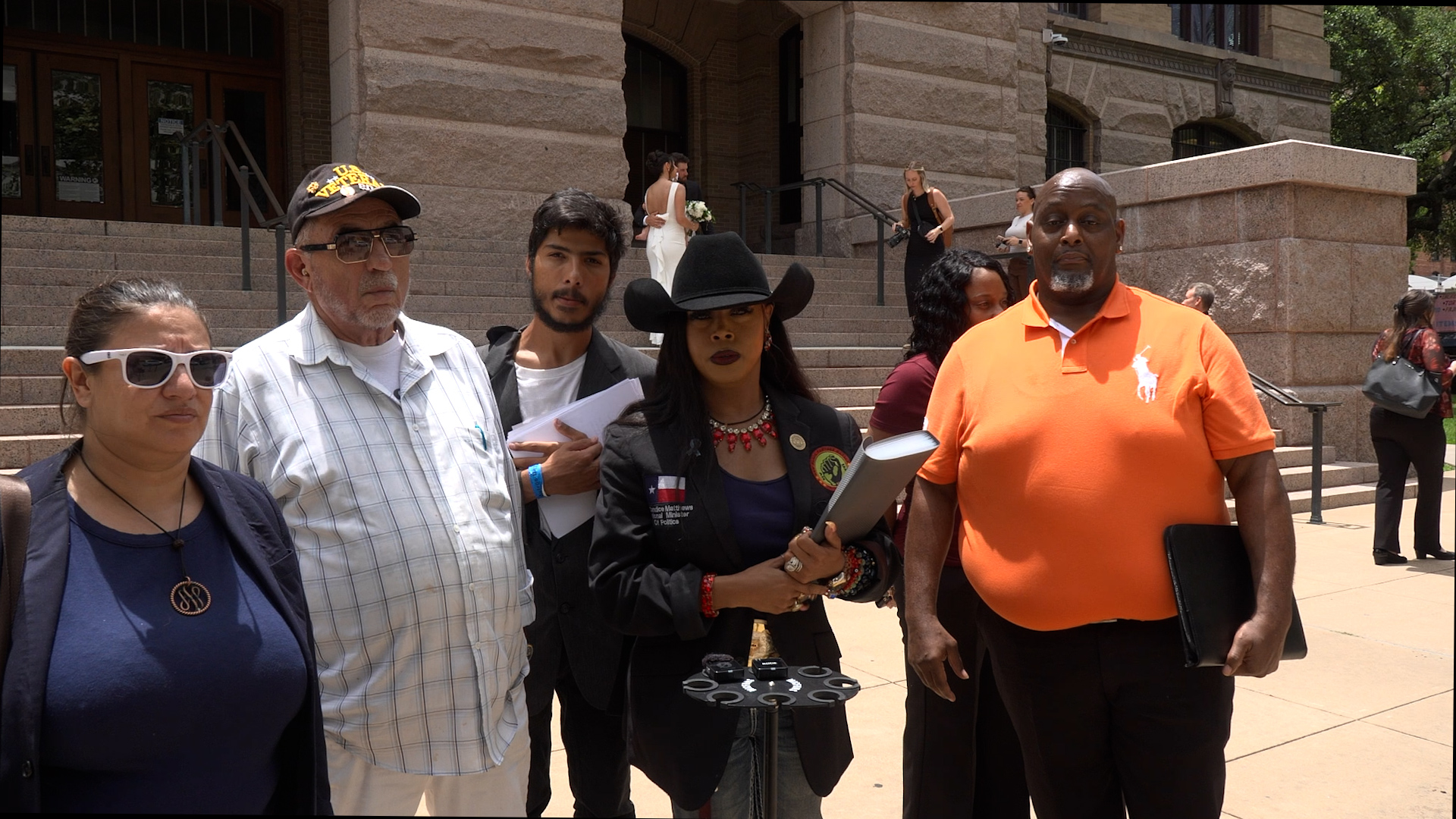 Group of diverse adults standing on courthouse steps outside a stone building, some holding papers and a planner, looking toward the camera during a public event.