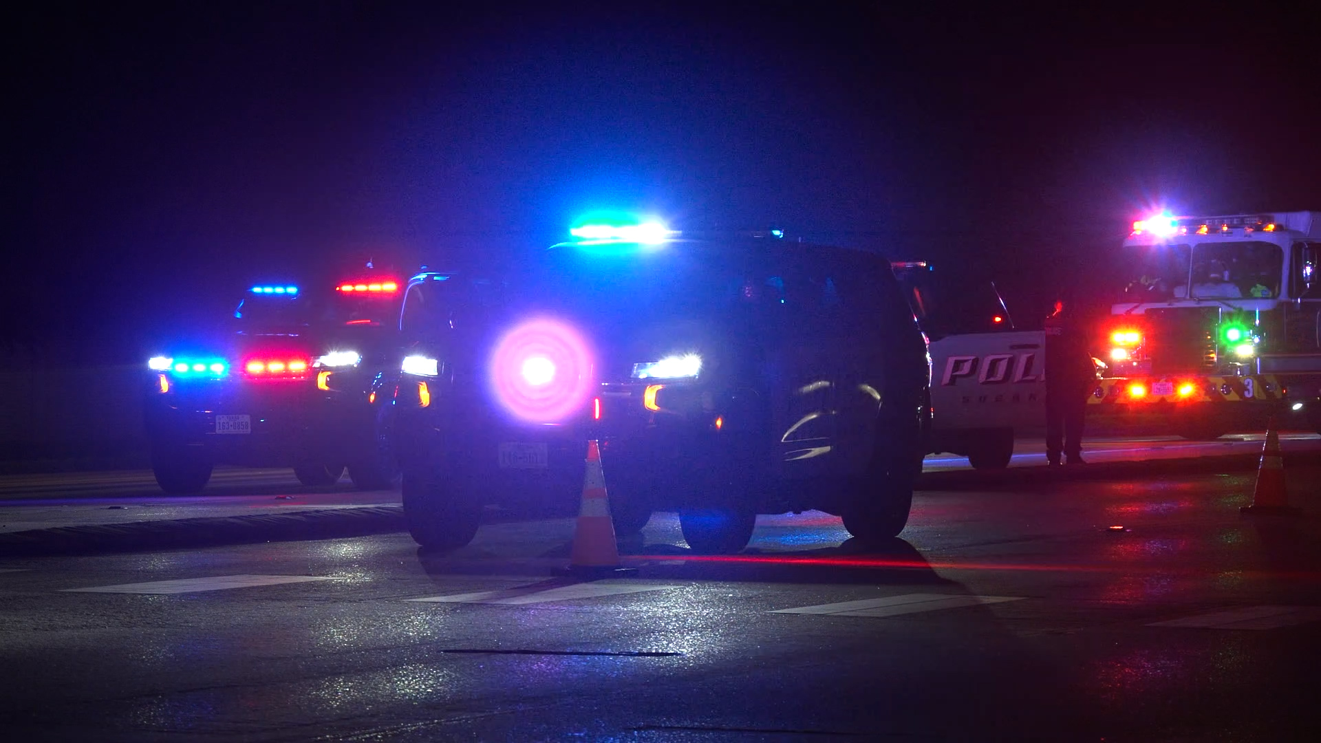 Nighttime scene of police cars and a fire truck with flashing red and blue lights on an empty street.