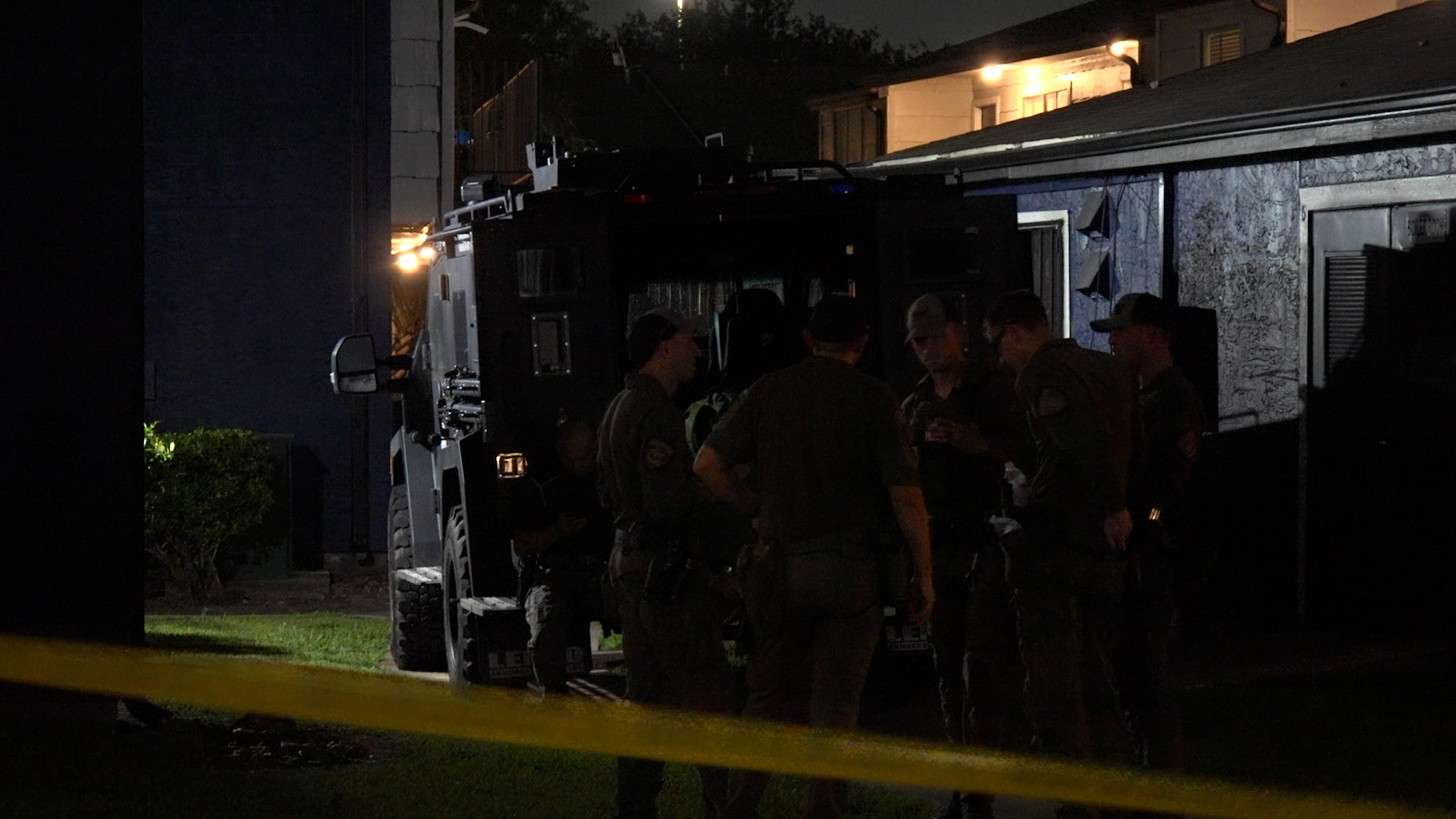 Nighttime scene with a group of uniformed officers gathered around a large armored vehicle behind yellow caution tape outside a building.