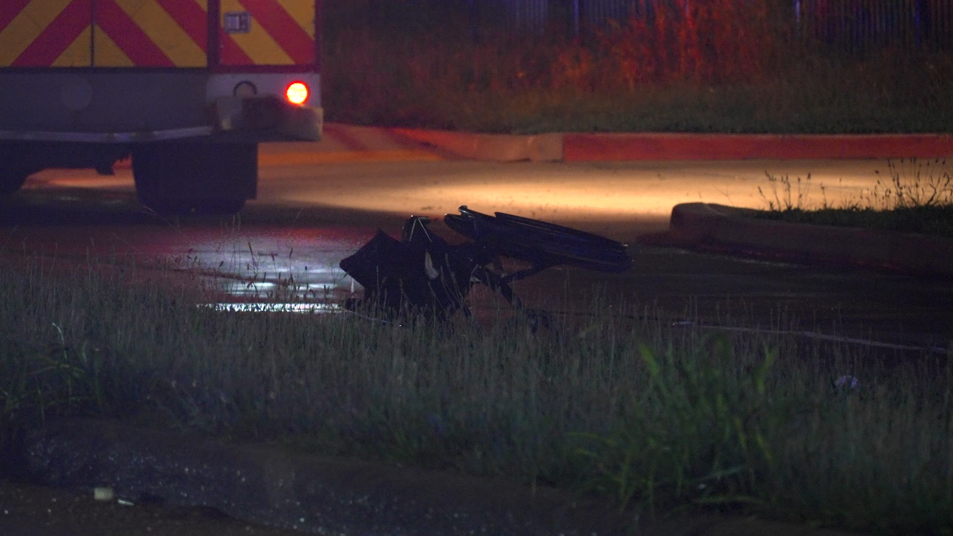 Night scene with a hazard-marked emergency vehicle on the left and scattered bent metal debris on a wet road at dusk/dark.